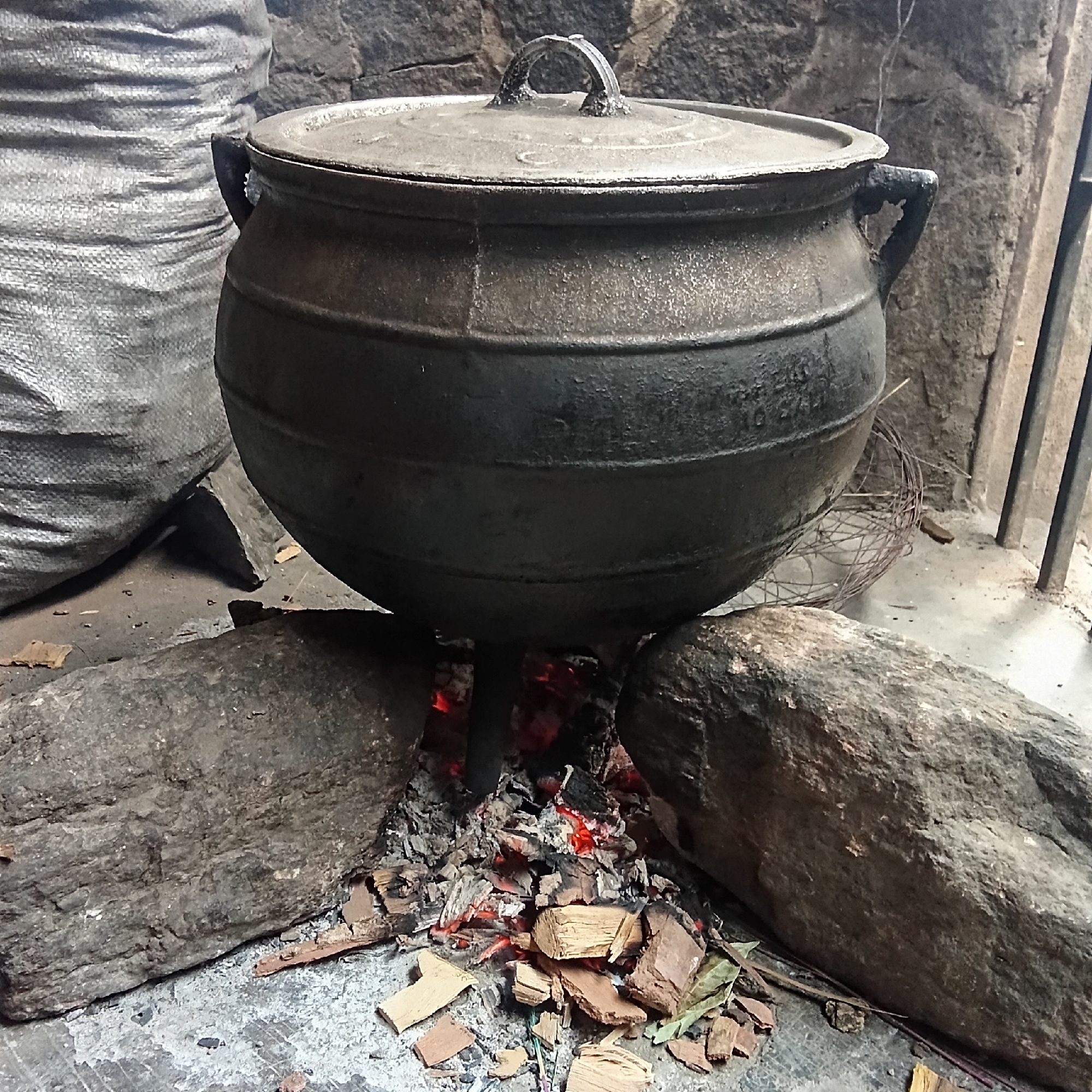 The image shows a large, dark, metal cooking pot with a lid, supported above a small fire. The fire is made of wood and embers, surrounded by three large rocks. The ground is a rough, gray surface, possibly concrete. In the background, there is a large, gray, woven bag, and a rough, stone wall. The location is Unknown, Unknown.