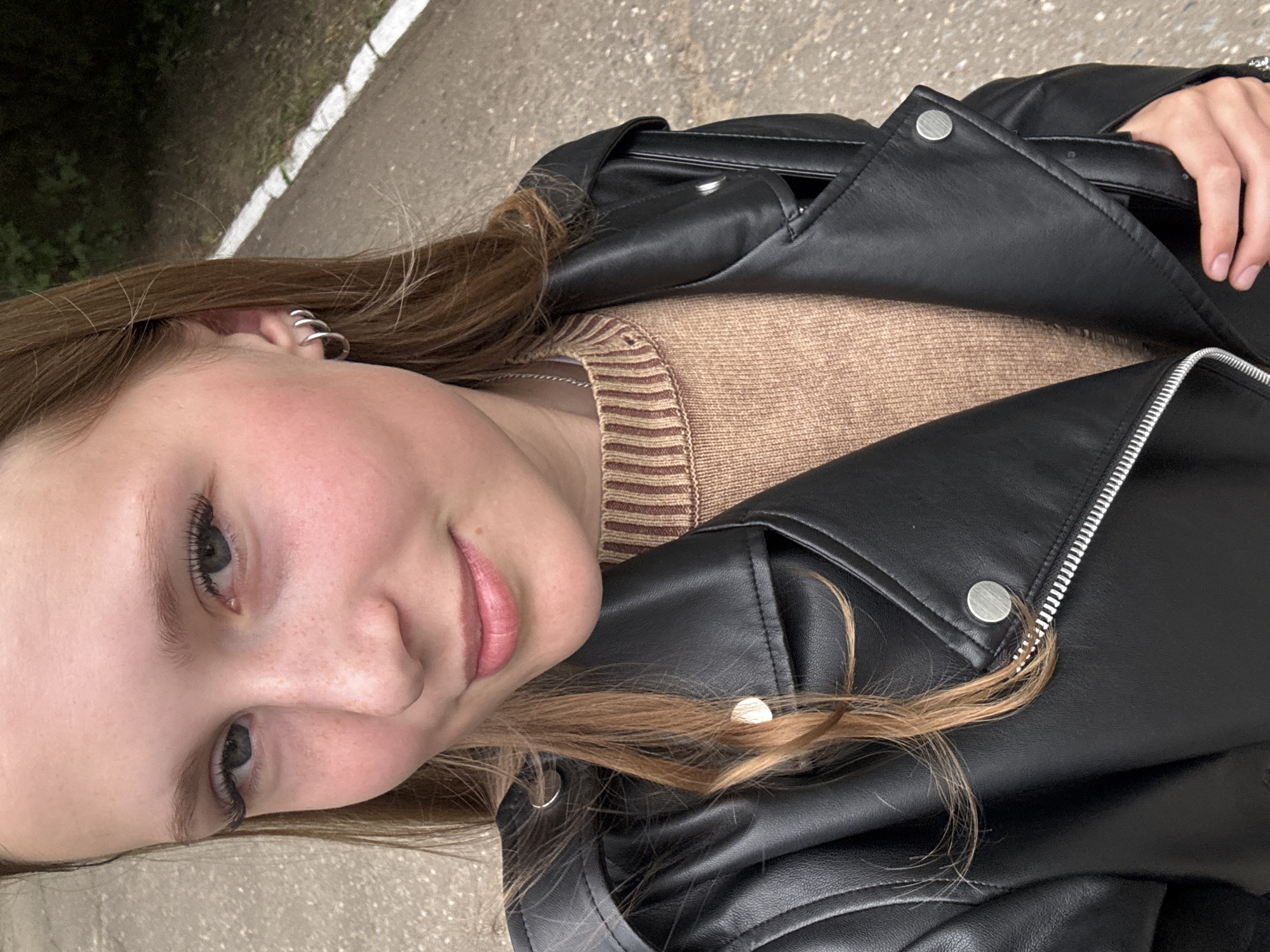 The media file is a close-up, rotated selfie of a young woman with long, light brown hair and fair skin. She is wearing a tan and brown striped sweater underneath a black leather jacket with a silver zipper and silver snap buttons. She has light blue eyes, visible freckles, and pink lipstick. She is wearing a silver hoop earring in her visible ear. The background appears to be an outdoor setting, possibly a pathway or road with some green foliage visible on the left. The lighting suggests it is daytime, and the weather appears to be overcast or perhaps it is just the lighting conditions of the photo. There is no visible text in the image. The woman has a relaxed and pleasant expression. The context provided places the location as Chaikovsky, Russia.