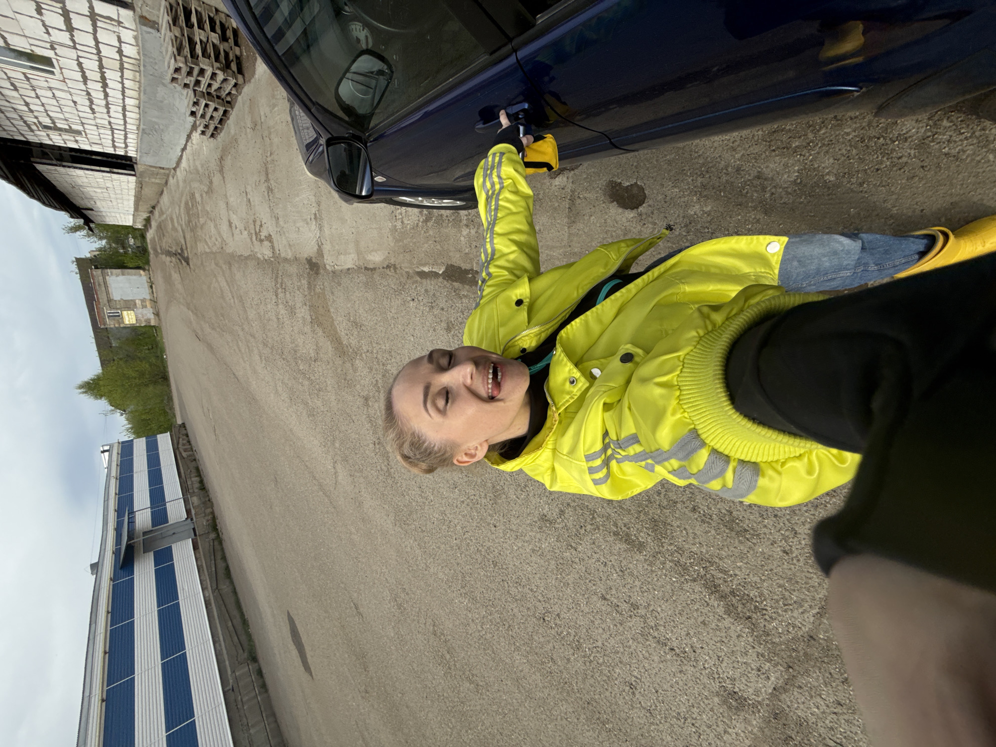 A woman in a yellow jacket and yellow boots is standing next to a blue car and appears to be opening the door. The scene takes place outdoors on an asphalt road next to a building with blue and white paneling. Another building with white bricks is in the background. The location is the city of Chaikovsky, Russia.