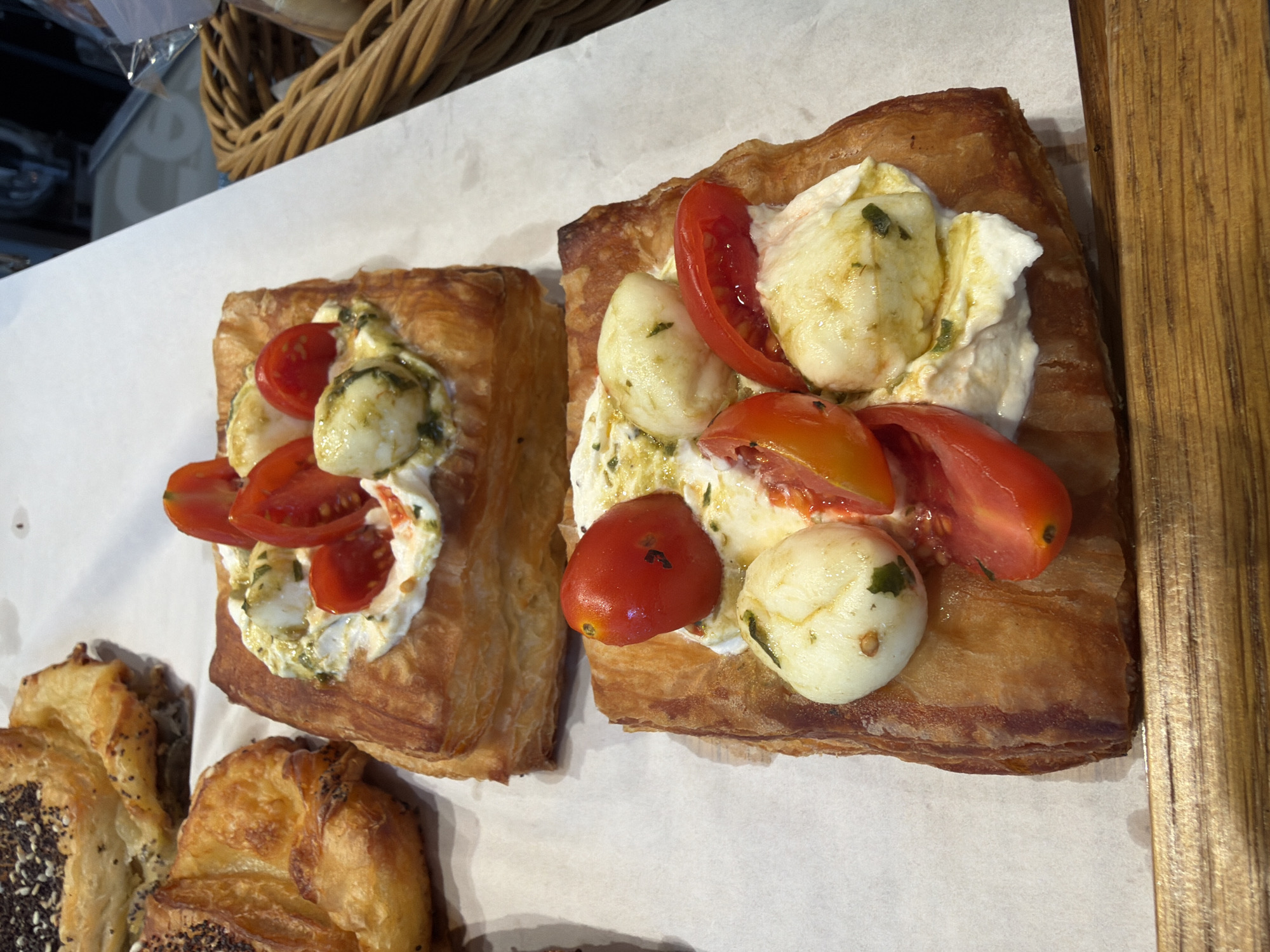 The image shows two pastries topped with mozzarella, tomatoes, and pesto on parchment paper. Part of a wicker basket is visible in the upper left corner and there is a wooden surface in the lower right. Additional pastries are partially visible in the lower left corner. The location is Unknown, Unknown.