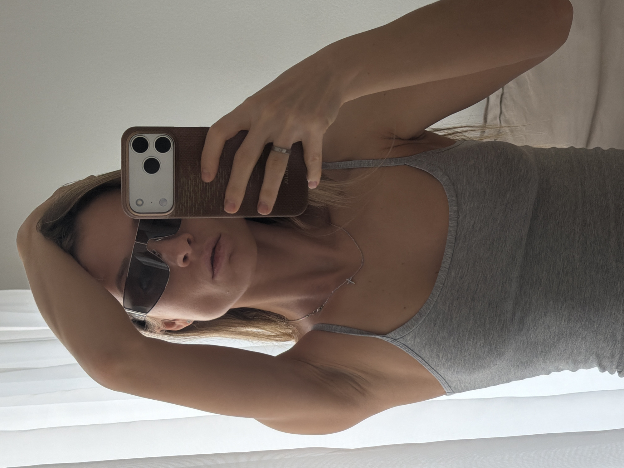 A person with light skin, wearing a gray tank top and sunglasses, is holding a phone with a brown case in front of their face, apparently taking a selfie. The person's left arm is raised and resting on their head. A silver ring is visible on their hand. A delicate silver necklace with a small cross pendant is around their neck. The background appears to be a white wall or curtain. There's no information to suggest the image was taken in Khimki, Russia.