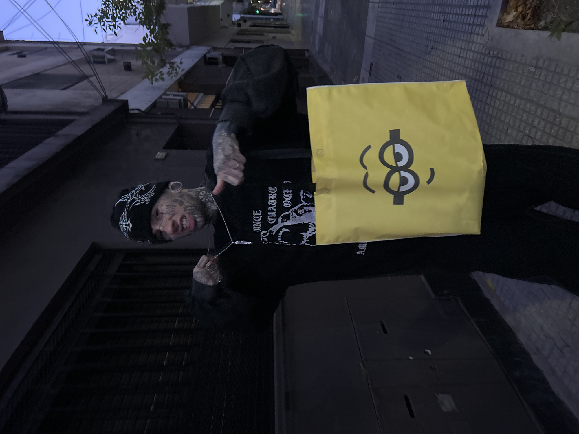 A person stands on a sidewalk next to a building in a narrow street in Buenos Aires, Argentina. The person, heavily tattooed on their face and hands, wears a black beanie and a black hoodie, displaying a silver necklace. They hold a yellow shopping bag with a black cartoon face graphic. The background reveals the street, sidewalk, and buildings on both sides, capturing an urban setting at dusk.