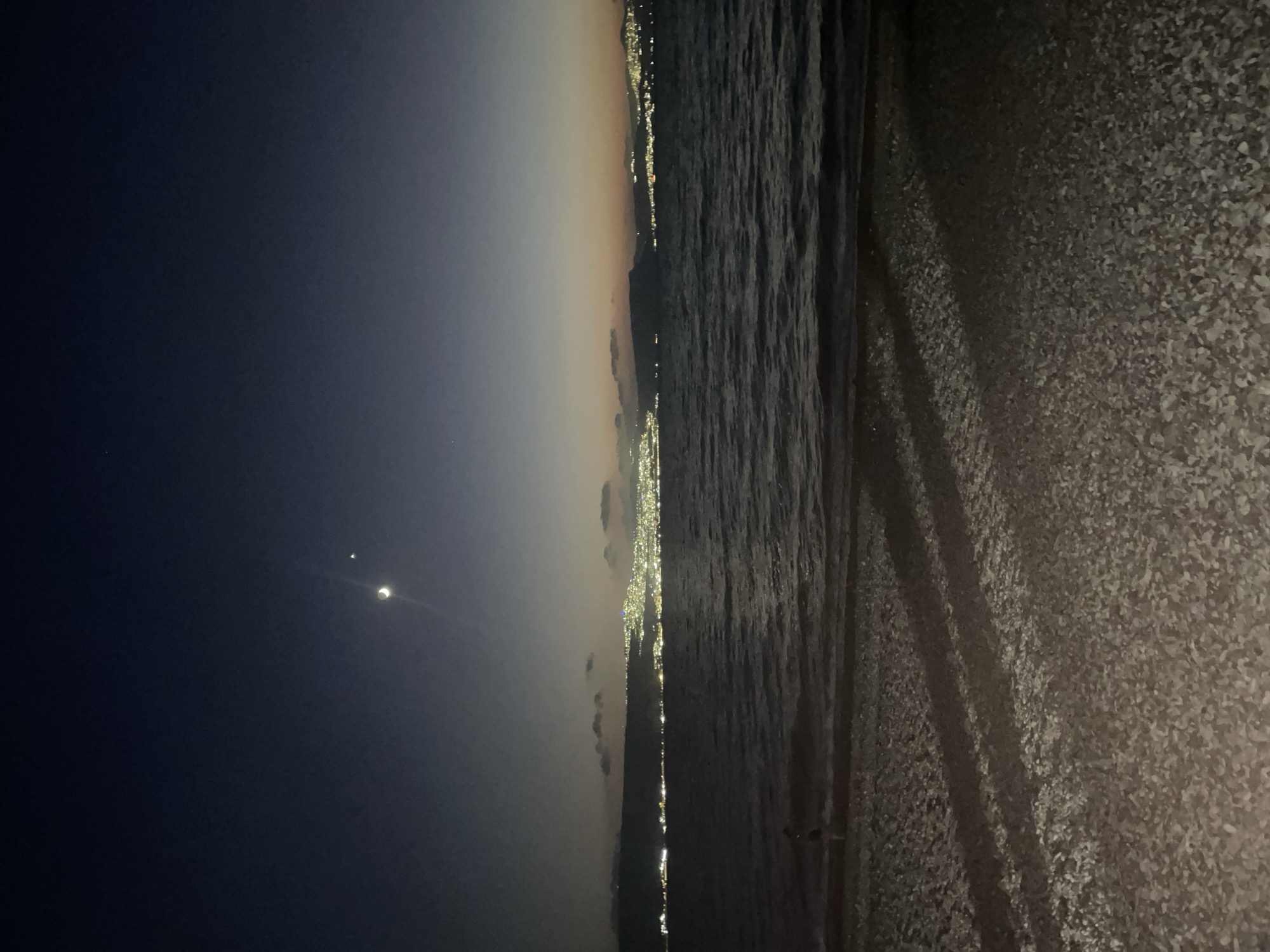 An image taken at night in the city of Emek HaYarden Regional Council, Israel, shows a serene seascape. The sky is a deep blue, transitioning to a lighter hue near the horizon where distant city lights glow. A crescent moon and a planet are visible in the upper sky. The sea is calm, with gentle waves lapping at a pebble beach in the foreground. The beach is illuminated by the city lights, casting long shadows across the stones. A solitary figure, possibly a child, stands near the water's edge. The overall atmosphere is peaceful and tranquil, highlighting the beauty of the night sky and the coastal landscape.
