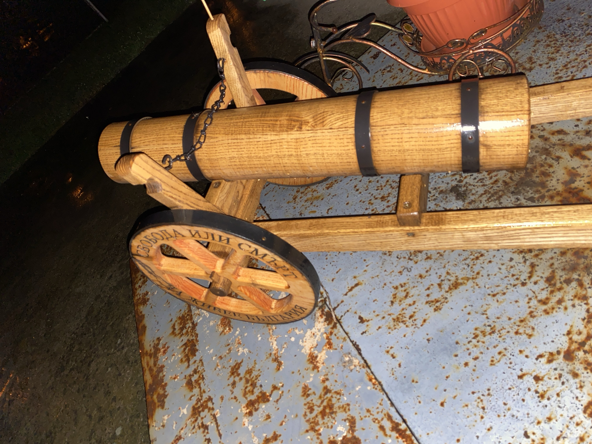 A wooden cannon sits on a rusted, light blue metal surface. The cannon has two black metal bands around the barrel and black wheels with the text "Свобода или смърт" (Freedom or Death) written in Cyrillic on the outer ring. The cannon is made of light brown wood and is chained to its support. To the right of the cannon, there is a wrought iron planter with a terracotta pot. The background shows a dark, blurry area that could be pavement or vegetation. The location is presumed to be Konstantinovo, Bulgaria based on the text.