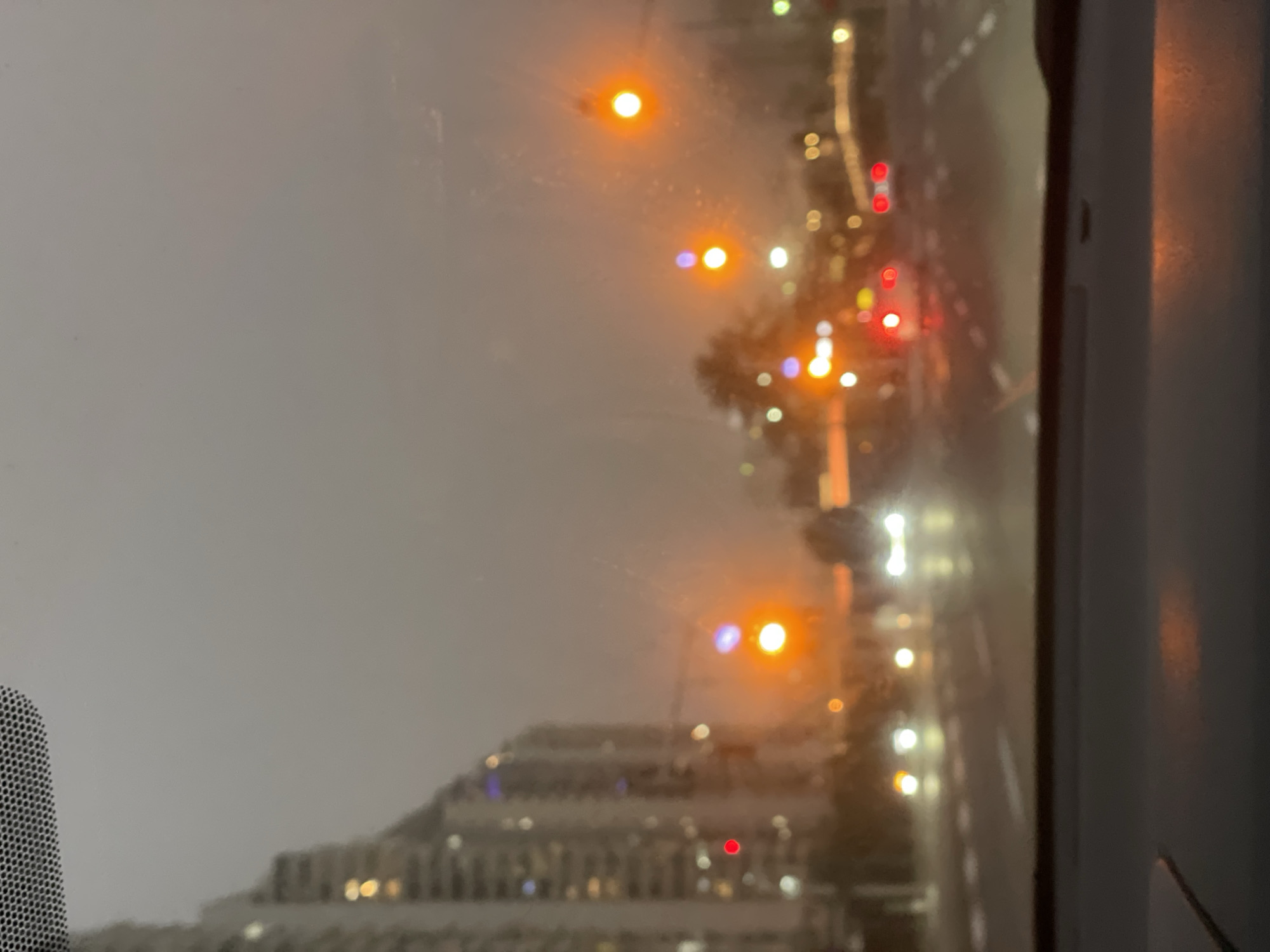 The image, taken from indoors, looks out onto an urban landscape on a cloudy or rainy night. Out-of-focus streetlights and traffic lights illuminate the blurred street scene. In the distance, a tiered building is visible. The location is Petah Tikva, Israel.