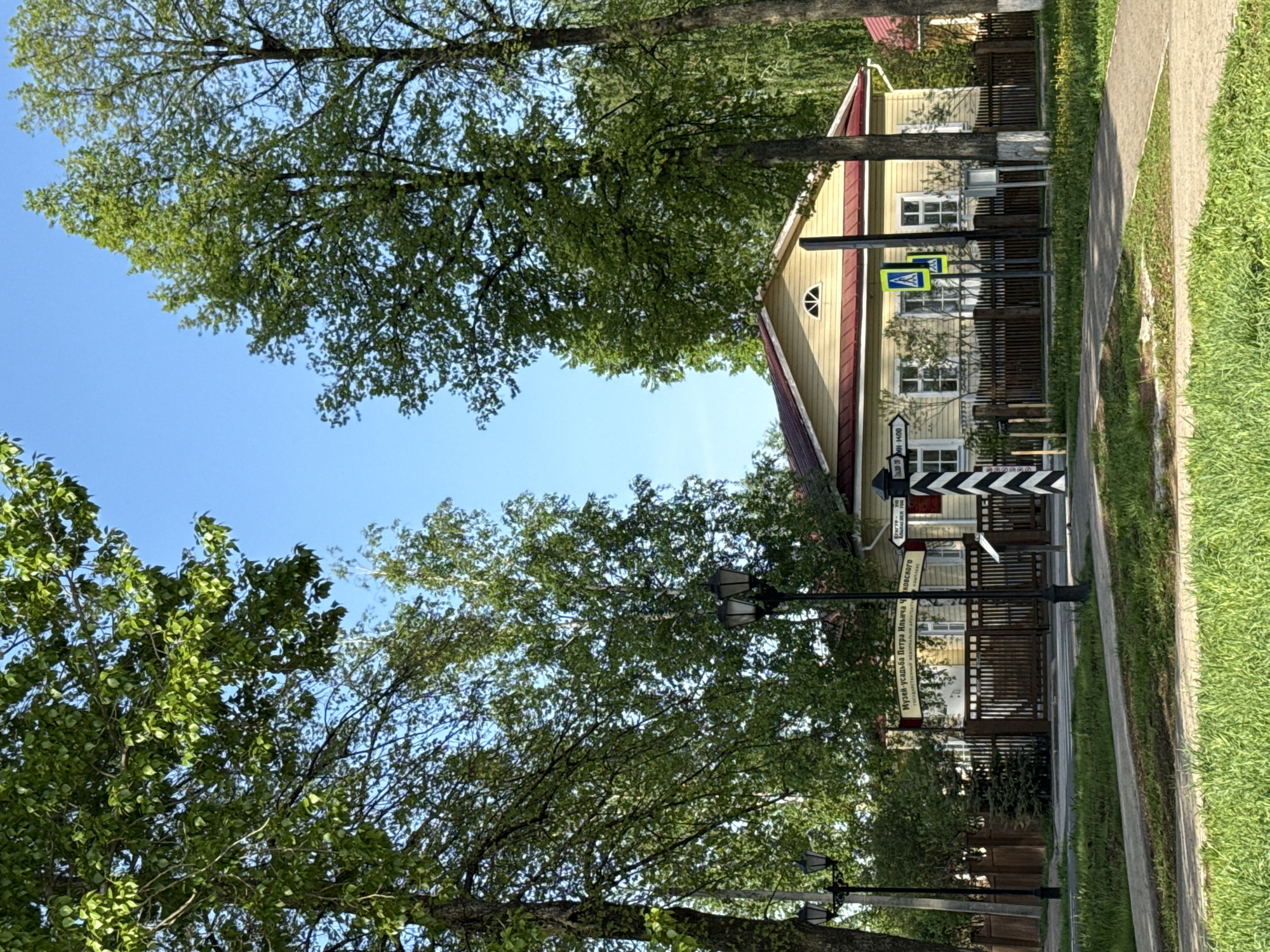 The image shows a street in Votkinsk, Russia, lined with trees and a sidewalk. A yellow house with a red roof is visible behind a wooden fence. Several street signs are present, including a pedestrian crossing sign and a directional sign with text in Russian, indicating destinations like "Museum-Estate of P.I. Tchaikovsky." Street lamps are visible along the street. The sky is blue with some cloud cover visible through the trees. The grass is green and neatly maintained.