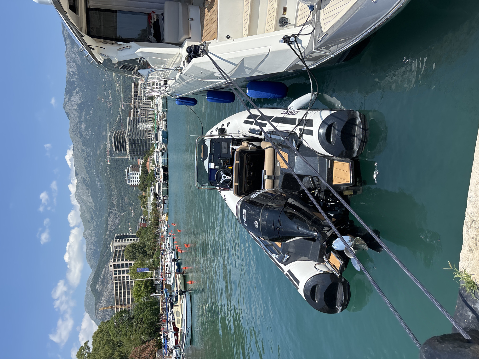 A tender is docked alongside a larger white yacht in a harbor with a city and mountains in the background. The tender is a rigid-hulled inflatable boat with black and white tubes and a black outboard motor. It is tied to the yacht with dark ropes. The yacht has a white hull and a teak deck. In the background, a city with tall buildings and cranes is visible, with a range of mountains behind it. The water is clear and blue. The sky is blue with scattered white clouds.