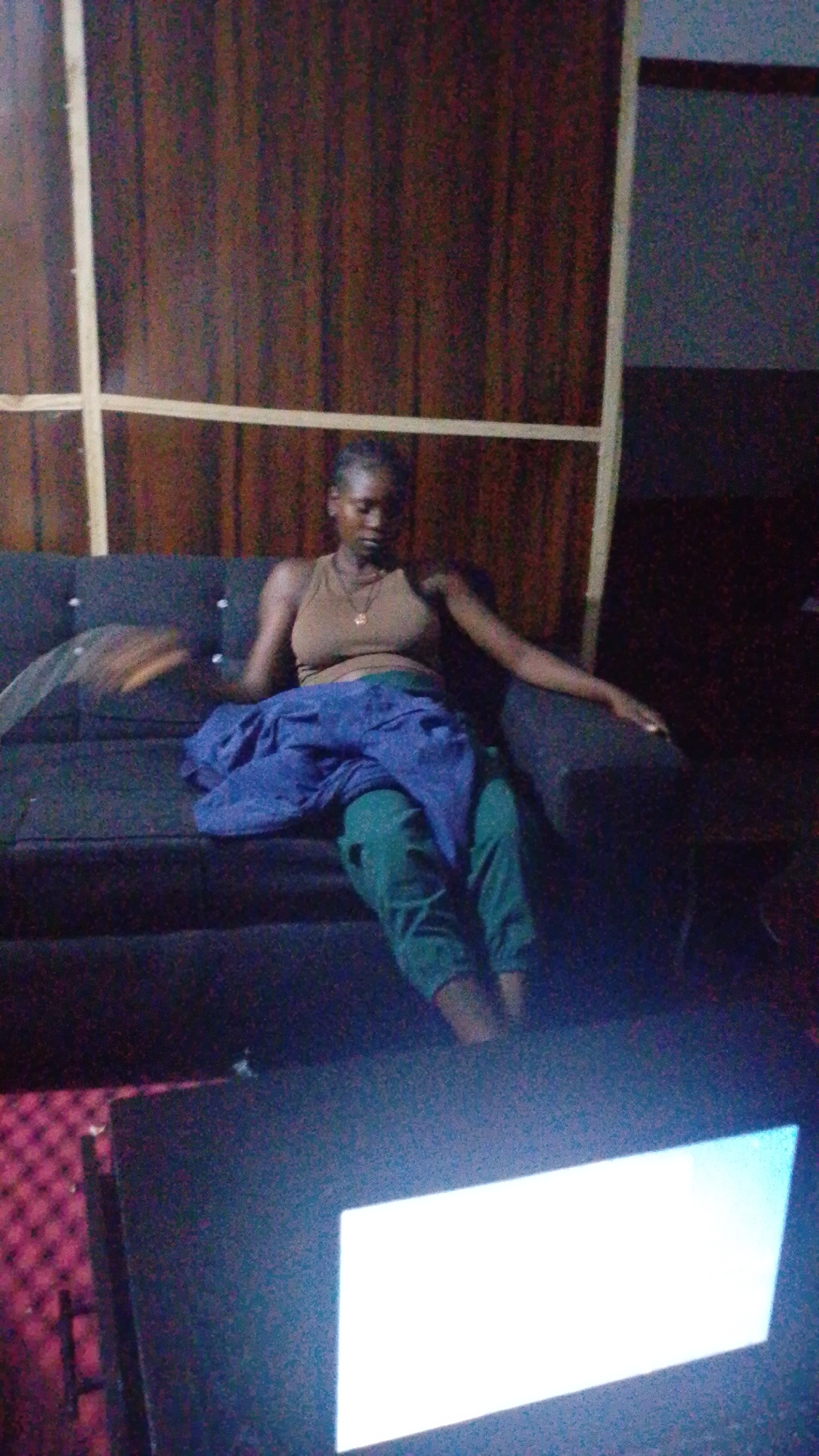 The image depicts a young woman seated on a dark-colored couch in what appears to be a dimly lit room. She is wearing a taupe tank top and green pants. A blue cloth is draped over her lap. Her posture suggests she is leaning back on the couch, with her left arm resting on the back of the couch. Her head is tilted down, and her gaze is directed towards her lap. The background features a dark, wood-paneled wall with vertical and horizontal light-colored beams. In the foreground, a portion of a surface with a red, checkered pattern is visible, and a bright, white rectangular light source is present, possibly from a screen. There are no clear indications of the specific location within Jos, Nigeria, but the indoor setting with the described elements provides a general context.