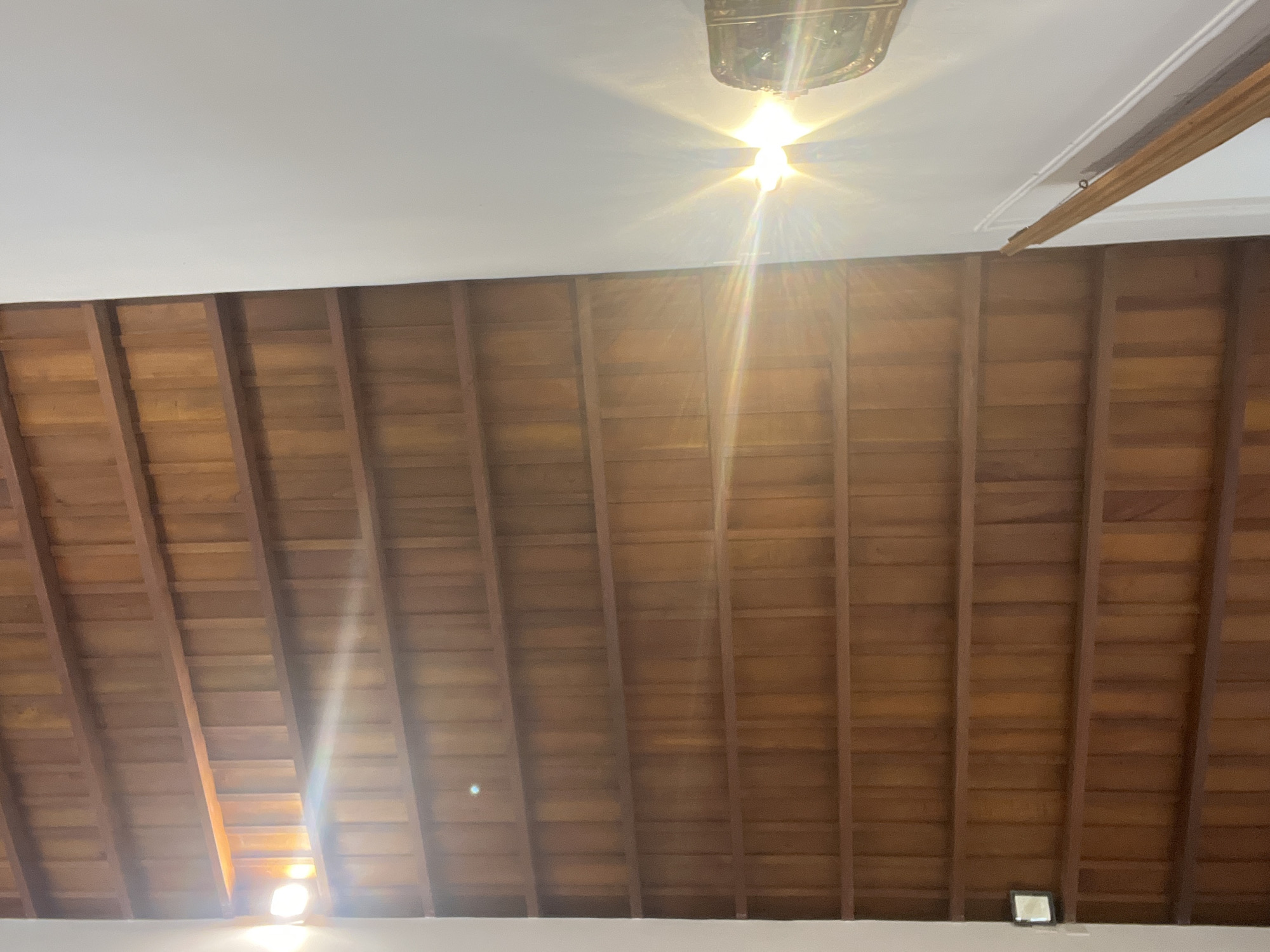 This image depicts an interior architectural space in Santa Ana, Venezuela, characterized by a prominent wooden slatted ceiling and contrasting white walls. The ceiling comprises numerous parallel horizontal beams with vertical wooden planks or slats set between them, forming a grid-like pattern of medium-brown wood.
On the right side of the image, a flat white wall occupies approximately one-third of the frame, extending diagonally from the upper right to the lower right. A wooden handrail is visible along the lower portion of this white wall, suggesting a staircase or elevated walkway. A strong, starburst-like light source emanates from a fixture or opening on this white wall, creating significant lens flare that extends across the ceiling. The fixture itself appears to be a decorative wall sconce, partially obscured by overexposure.
On the far left, a vertical white structural element, likely a wall or column, borders the wooden ceiling. Another bright light source is visible behind or adjacent to this structure, also producing a strong starburst effect and a horizontal beam of light that traverses the upper-left portion of the wooden ceiling. In the bottom-left corner, a small, dark, square object with a reflective surface is mounted on the white vertical element. Scattered across the wooden ceiling, several small, circular, blurry blue-green specks are present, likely reflections of dust particles or lens artifacts. No people or specific actions are depicted.