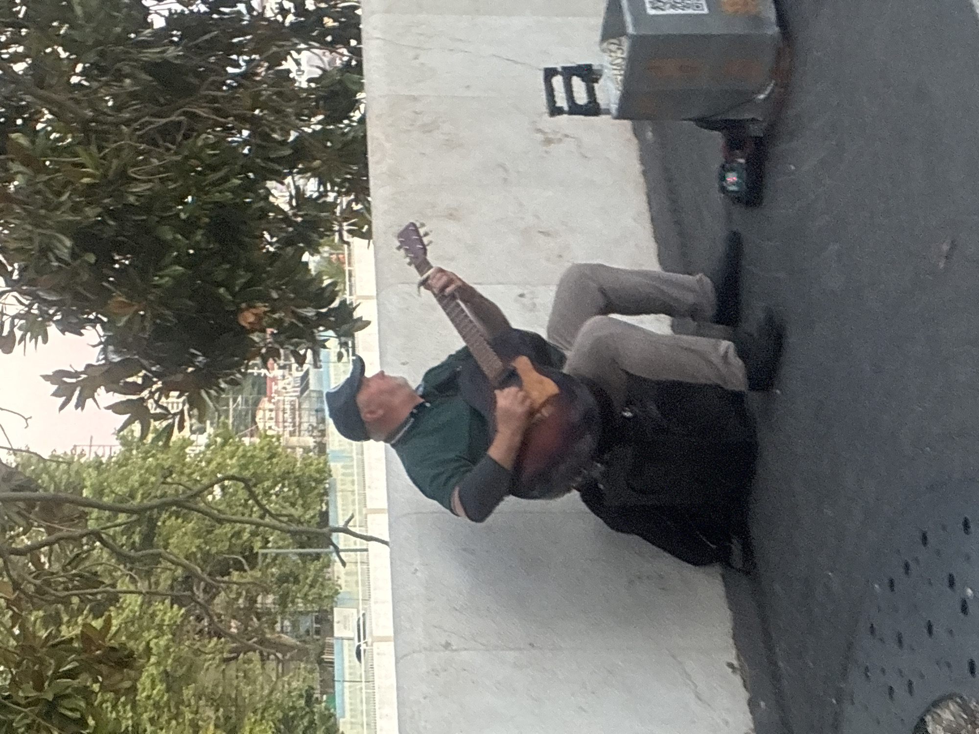 A man is playing an acoustic guitar while sitting on a black bag. He is wearing a dark blue baseball cap, a green collared shirt with dark stripes on the sleeves, and light brown pants. The man is seated on a dark, textured surface, possibly asphalt, with a light-colored concrete wall behind him. Trees with green leaves are visible in the background, with some buildings and what appears to be a crane in the distance, suggesting an urban environment. To the right of the man, there is a metal container with some items inside, and a small electronic device on the ground in front of it. The scene is illuminated by natural daylight.