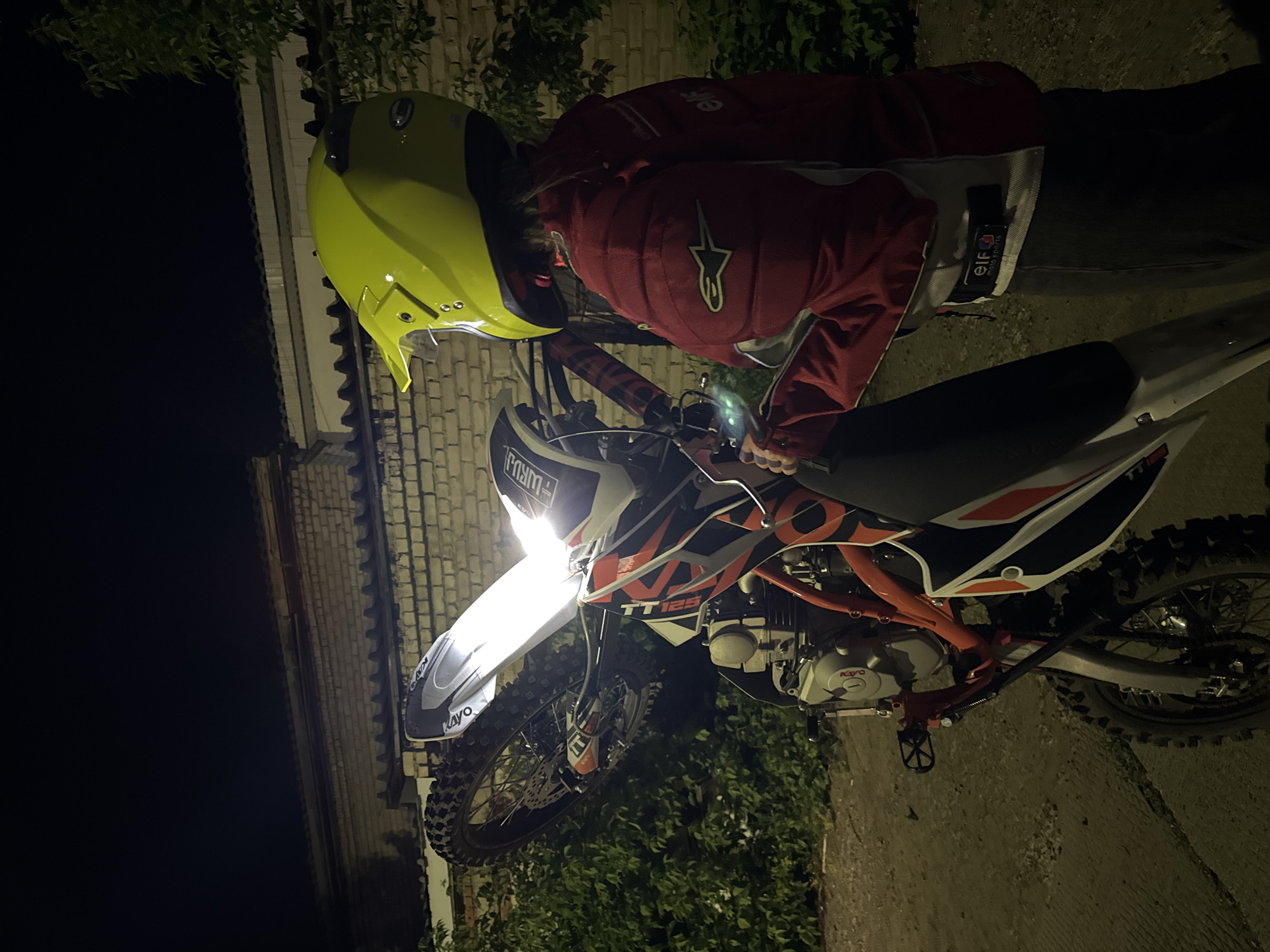A person wearing a yellow helmet and red jacket stands next to a Kayo TT125 dirt bike at night. The bike's headlight is on. The location appears to be near a brick building in Chaikovsky, Russia, based on the style of the building and surrounding vegetation. The area is dimly lit. The person is looking down at the bike.