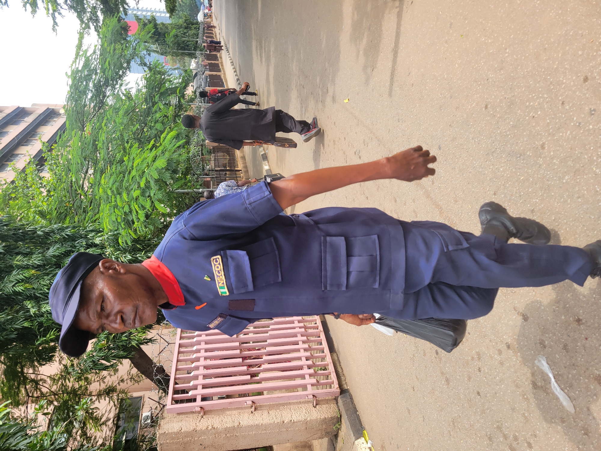 The image captures an outdoor urban scene in Abuja, Nigeria, during the daytime. The primary subject is a man in the foreground, wearing a navy blue uniform with a red collar and a matching cap. His uniform features several pockets, and on his left chest pocket, an emblem with the visible text "NSCDC" is displayed, indicating he is likely associated with the Nigeria Security and Civil Defence Corps. He appears to be holding a black bag in his left hand, and his right arm is slightly extended.
In the mid-ground, several other individuals are walking away from the camera on a light-colored paved road. One person in a dark shirt and trousers is particularly noticeable. To the left of the frame, a multi-story building with numerous windows is visible. The scene is rich with greenery, featuring lush trees and bushes lining the street, creating some shade. A pink metal fence, topped with what appears to be barbed wire, runs parallel to the street on the left. Further in the background, more buildings, including some with modern, reflective surfaces, can be seen under a bright, sunny sky. The overall lighting suggests a clear day.