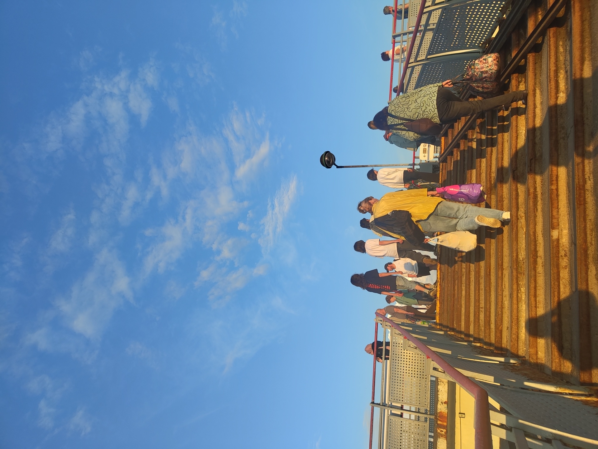 A group of people are ascending a weathered, outdoor staircase under a clear blue sky with scattered white clouds. The staircase appears to be made of metal with rust visible on the steps. Some individuals are carrying bags and packages. The scene is likely located in the city of Ramenskoye, Russia, given the presence of a distinctive street lamp at the top of the stairs. The perspective is from a low angle, looking up the stairs towards the top where the light is situated. The sunlight casts long shadows on the steps, indicating it is either early morning or late afternoon.