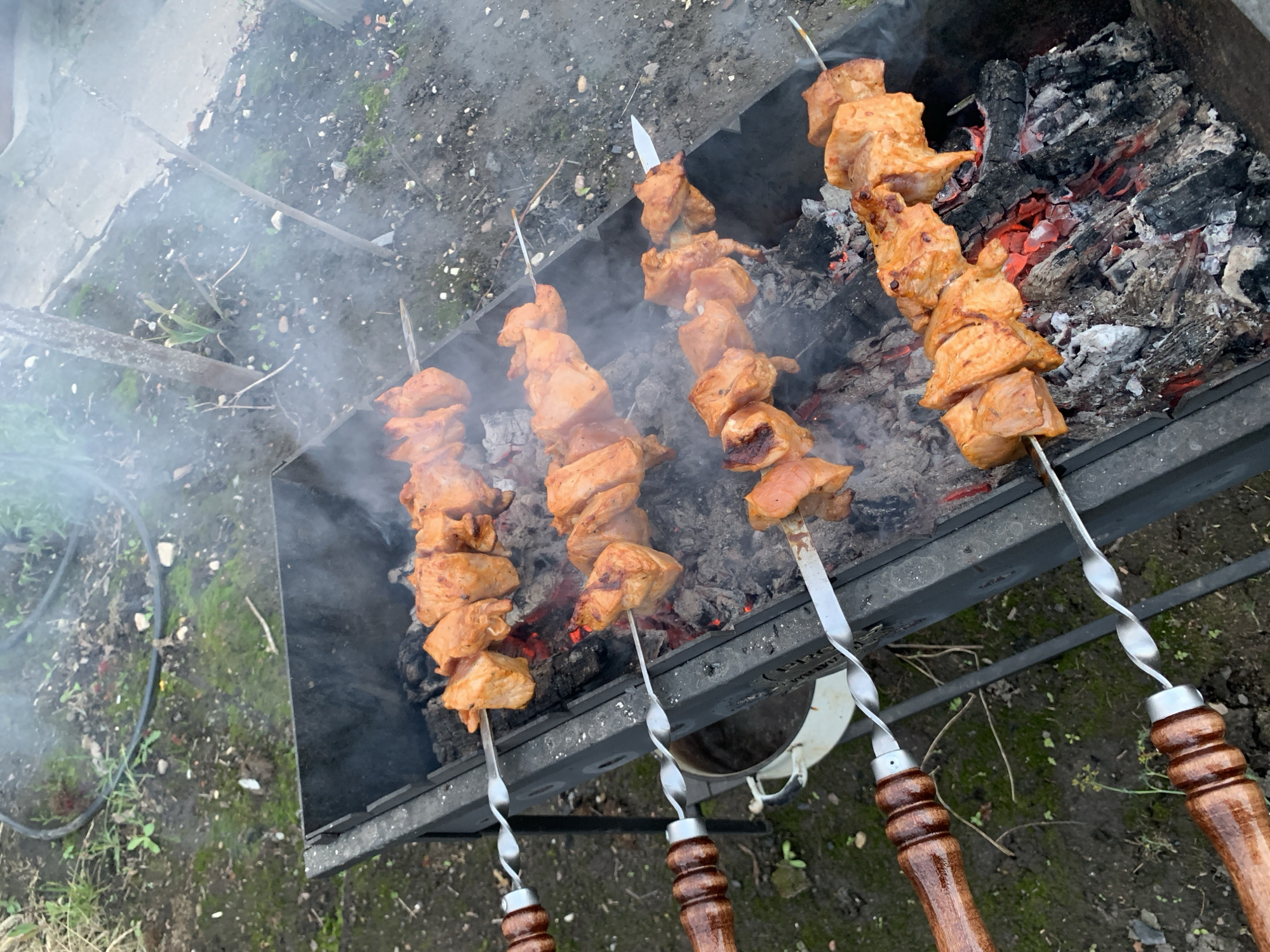 The image depicts four skewers of raw, marinated meat being grilled over hot coals in an outdoor barbecue. The meat appears to be chicken or pork, cut into bite-sized pieces and threaded onto metal skewers. The skewers are placed horizontally across a black metal grill, with some smoke rising from the coals. The environment is an outdoor setting, with dirt, grass, and small rocks visible on the ground surrounding the grill. In the background, a white bucket and what appears to be a hose are present. The lighting suggests an overcast or shaded day. The scene is consistent with a casual outdoor cooking event in a rural or suburban setting. The location is specified as Блеўчыцкі сельскі Савет, Belarus.