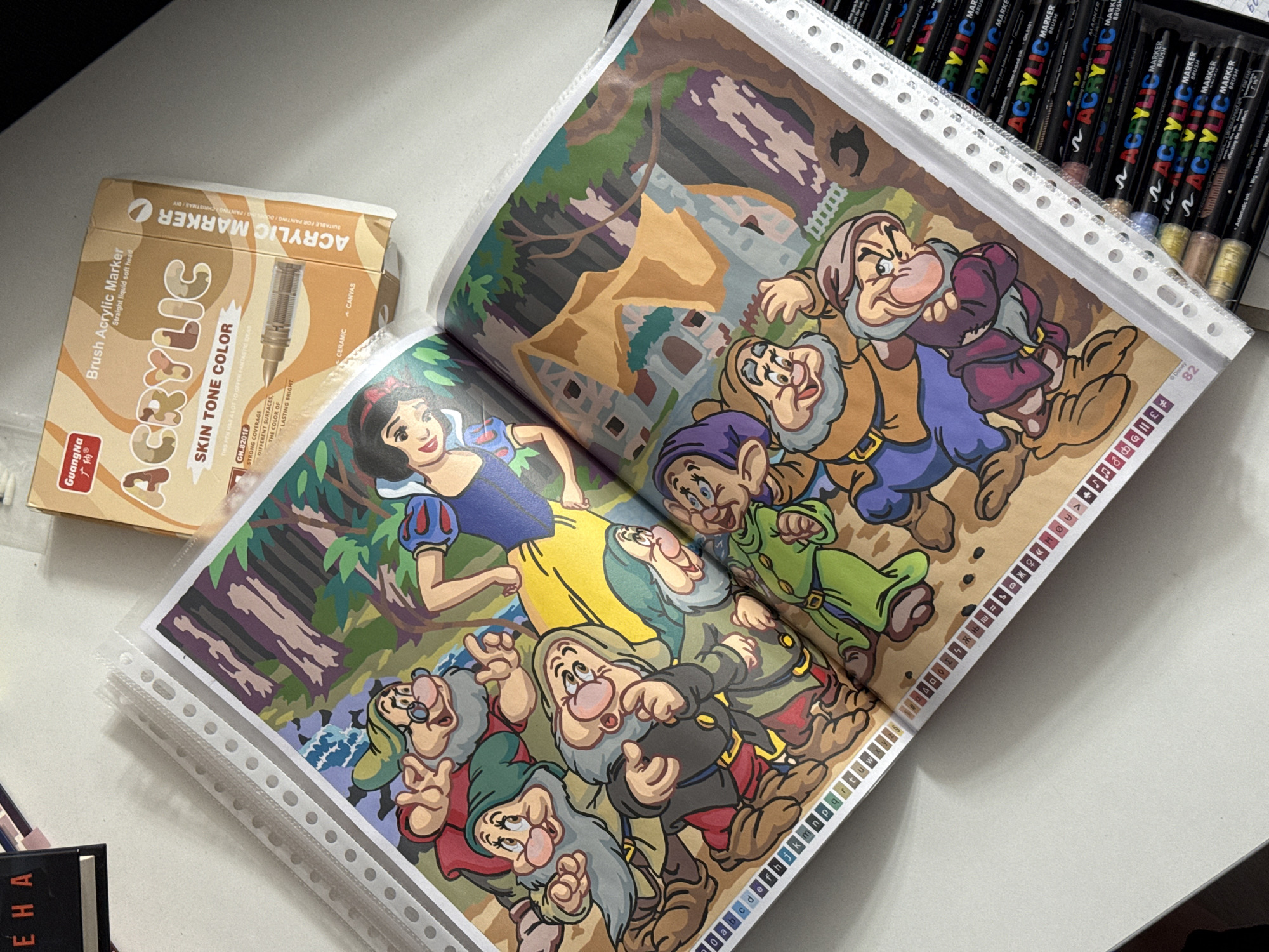 The image shows an open book, likely a coloring book, featuring an illustration of Snow White and the Seven Dwarfs. The book is placed on a white surface, possibly a table, next to a box of "GuangNa Acrylic Marker" pens, specifically the "Skin Tone Color" set. The book is protected by a clear plastic page protector. The illustration is in progress, with some areas colored and others left blank. The bottom edge of the book displays a legend of color codes, indicated by letters and symbols. Several pens are also visible in the lower right corner of the image, stacked in a container, with the brand "ACRYLIC MARKER" clearly visible on their labels. The overall scene suggests an art or craft activity. There is no visible indication of the city of Łódź, Poland, or any specific urban environment.