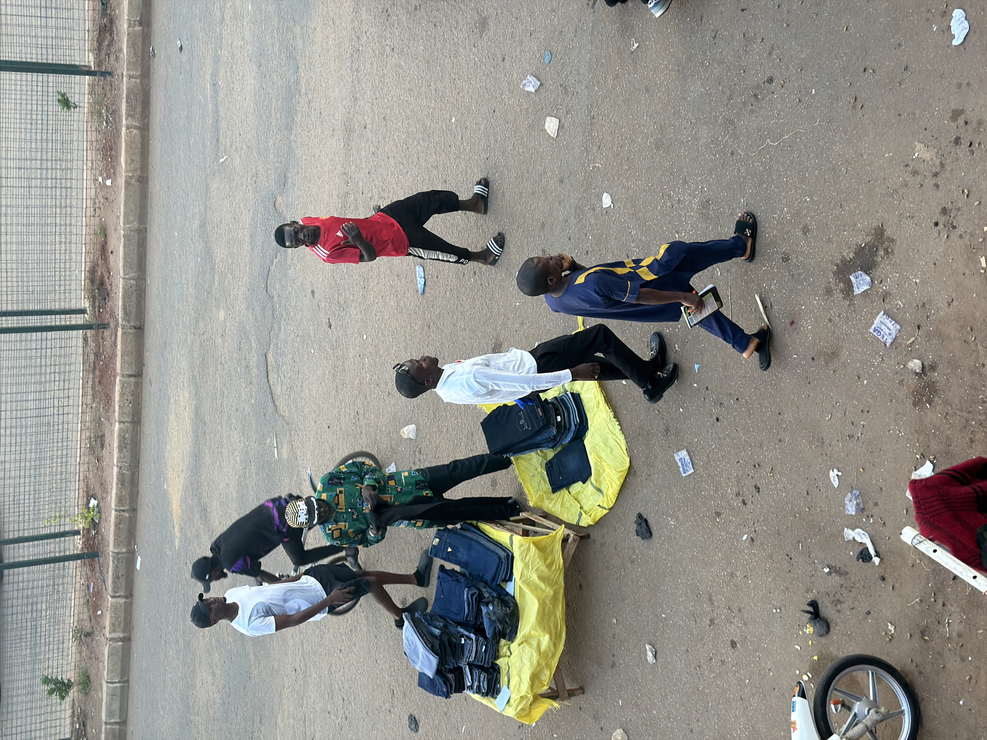In Bukuru, Nigeria, on a paved road with some debris scattered, six men are present. One man in a white t-shirt and black shorts stands looking to his left. Another man in a black shirt with purple accents rides a bicycle. A third man, wearing a colorful green and black shirt and black pants, is examining some dark fabric in his hands. A fourth man in a red t-shirt and black pants stands with his hand on his chest, looking to his right. A fifth man in a white long-sleeved shirt and black pants walks towards the right. The sixth man, wearing a blue and yellow patterned tunic and dark pants, walks to the left holding a small black object in his hand. In the foreground, a white motorcycle is visible with its front wheel turned to the right. A pile of blue jeans is displayed on a yellow tarp on a wooden structure. Another pile of dark pants is on a yellow tarp on the ground to the right. A metal railing with vertical posts runs along the top of the image, with a patch of dry grass and dirt behind it.