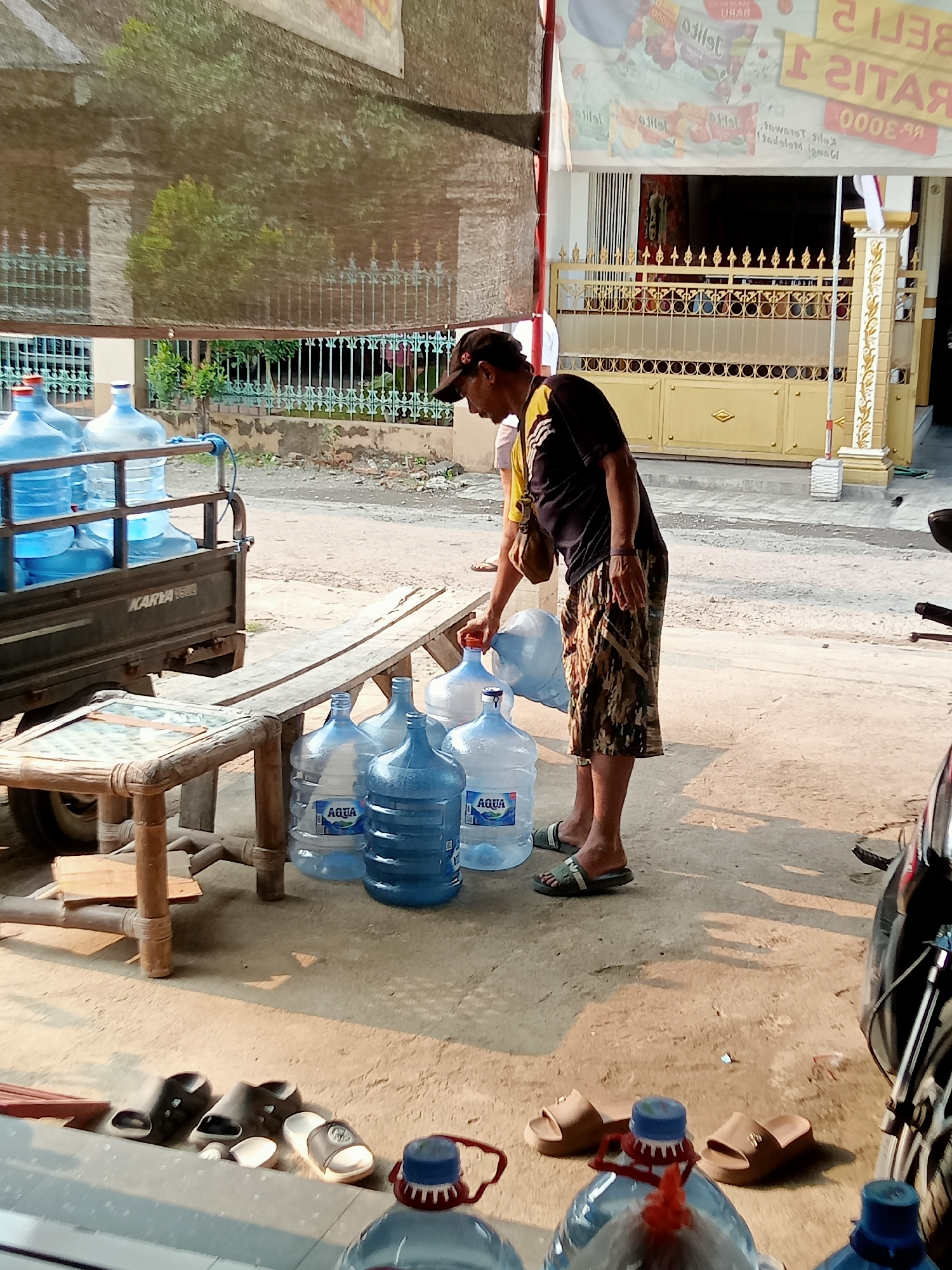 In Simo, Indonesia, a man wearing a dark t-shirt with yellow accents and camouflage shorts is shown on a sun-drenched street. He is handling large, blue water bottles, likely for delivery or refilling. Several of these bottles are lined up on the ground, with a few more stacked in the back of a small, flatbed utility vehicle labeled "KARYA 500". The vehicle appears to be a type of cart or trailer. Adjacent to the bottles on the ground is a wooden bench or table, also appearing to be made of rustic materials. In the background, a golden gate with decorative elements and a building with signage are visible. The ground is a mix of dirt and concrete. Several pairs of sandals are scattered near the foreground, suggesting a casual or work environment. The overall scene depicts a typical street-level transaction or delivery of bottled water in a commercial or residential area.