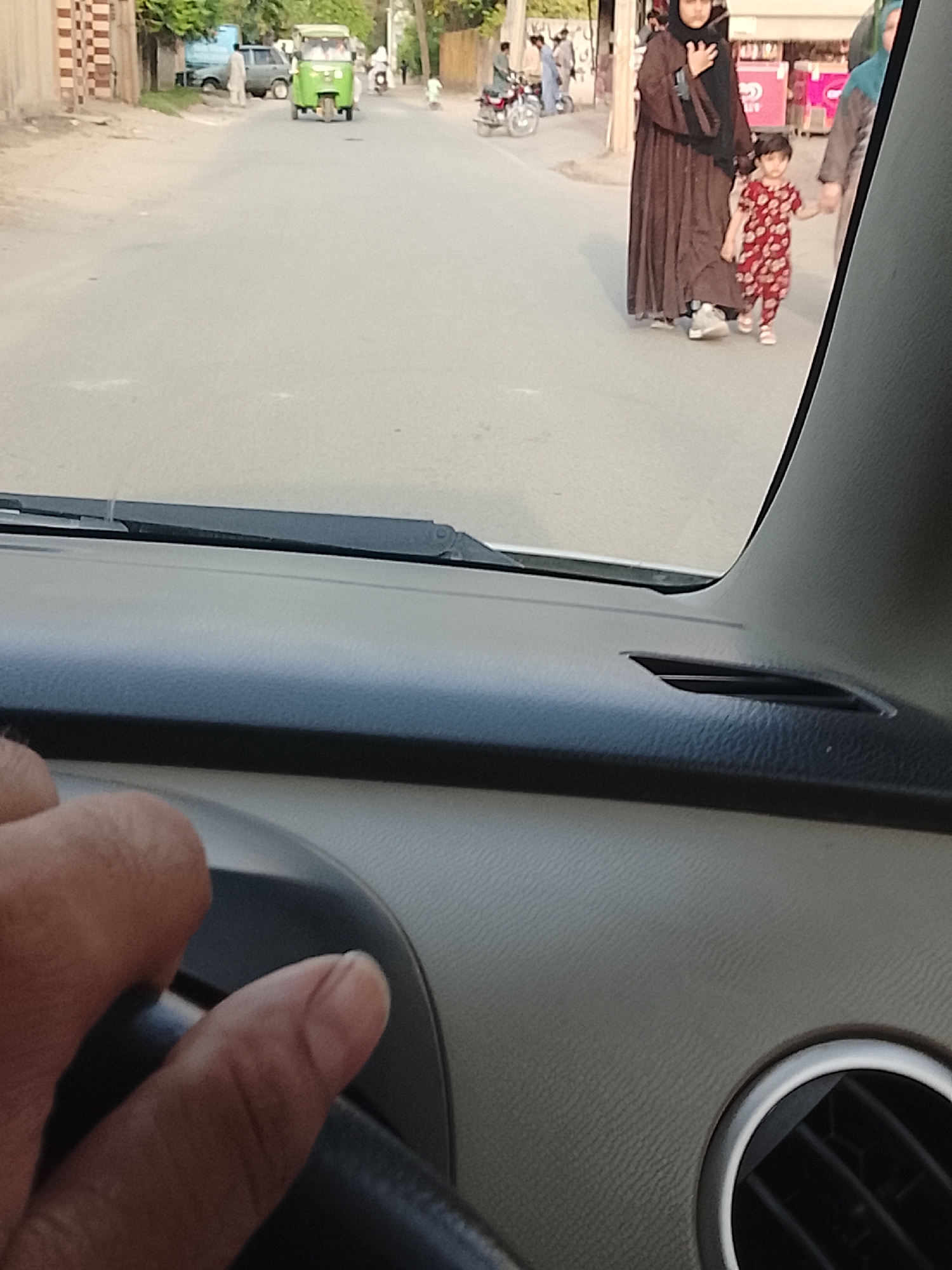 The scene, viewed from inside a car, shows a street in Nasheman Colony, Pakistan. A woman in a brown abaya and black hijab is walking along the side of the road, holding hands with a child wearing a red and white patterned outfit. Other individuals are visible further down the street, along with a green auto-rickshaw and parked motorcycles. The car's dashboard and steering wheel are in the foreground, suggesting the photo was taken by the driver.