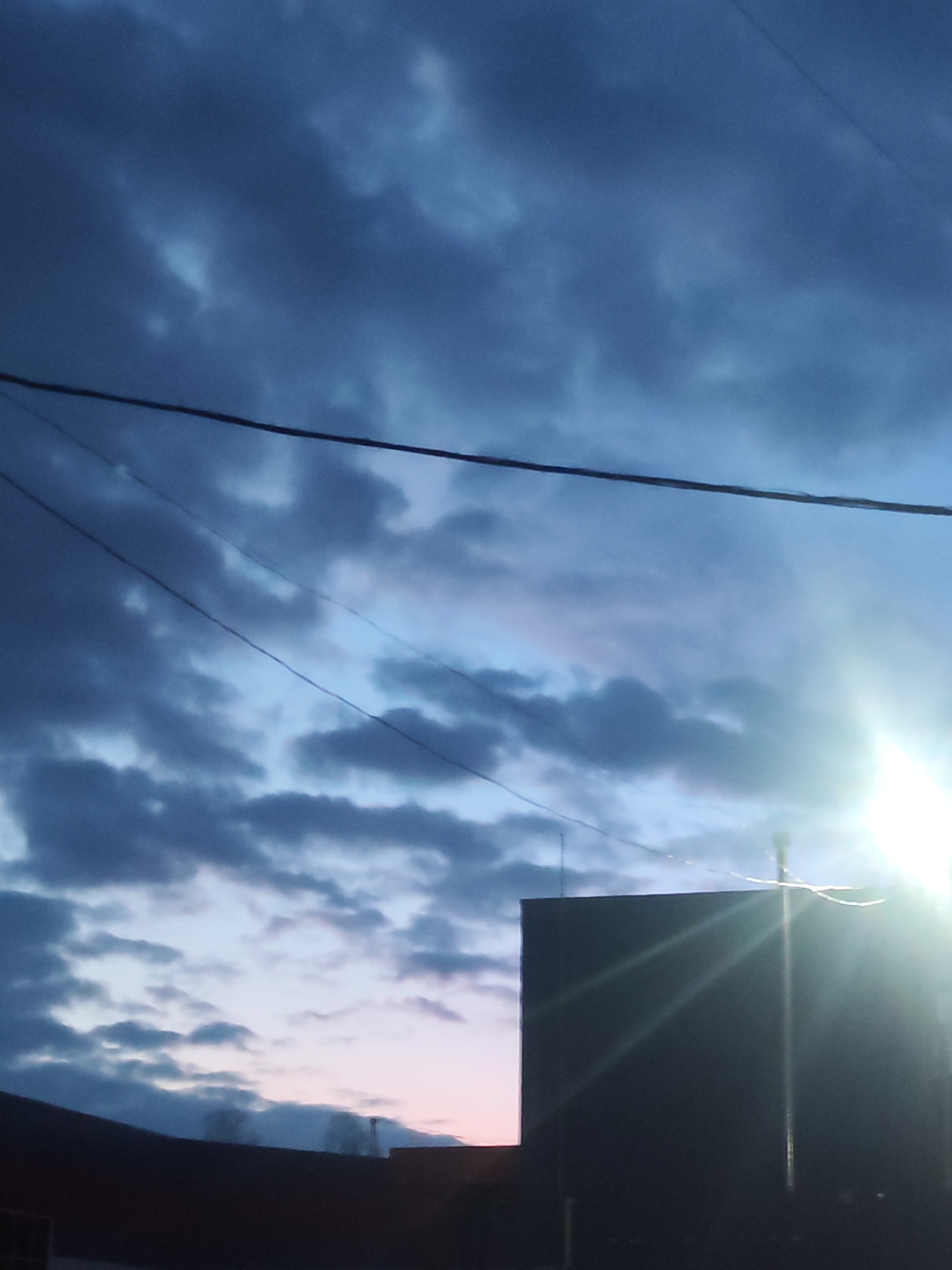 The image shows a twilight sky over Chaikovsky, Russia. The sky is predominantly blue with patches of dark, layered clouds. Power lines stretch across the upper portion of the frame. In the lower portion of the image, the silhouette of a building stands in the foreground, with a chimney rising beside it. A bright light source, possibly the sun or street light, is visible to the right of the building, creating lens flare and obscuring details. The buildings appear to be of modest height, likely residential or small commercial structures. The general environment conveys an urban setting during twilight or early morning.
