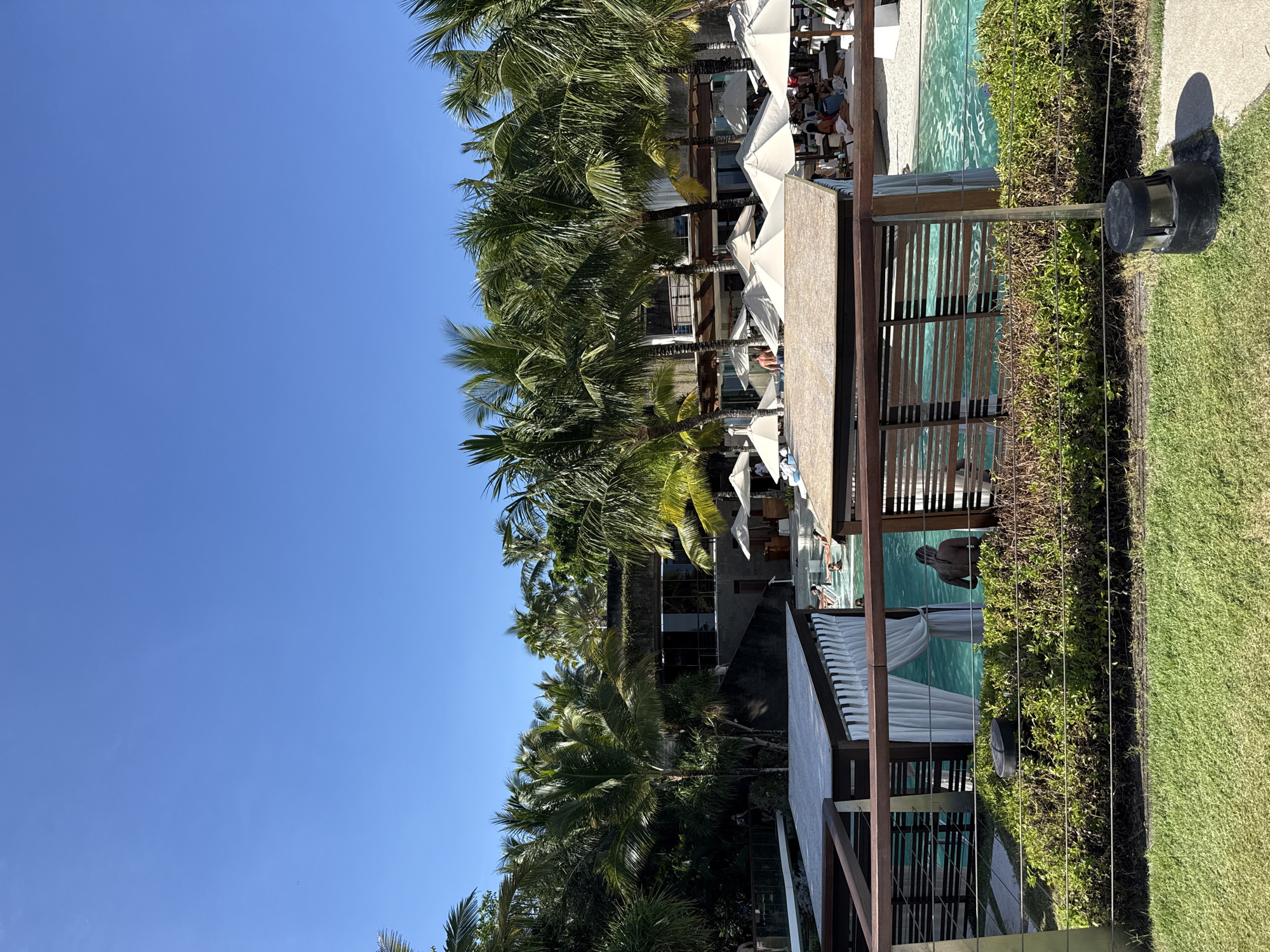The image shows an outdoor pool area, likely within a resort or hotel in Kuta Selatan, Indonesia. Several palm trees are visible, as is a clear blue sky. The pool is surrounded by lounge chairs and white sun umbrellas. A wooden cabana structure sits along the edge of the pool, with people in the water. Adjacent to the pool is a grass lawn. A dark, cylindrical object, perhaps a light fixture, is positioned on the grass near a hedge or fence.