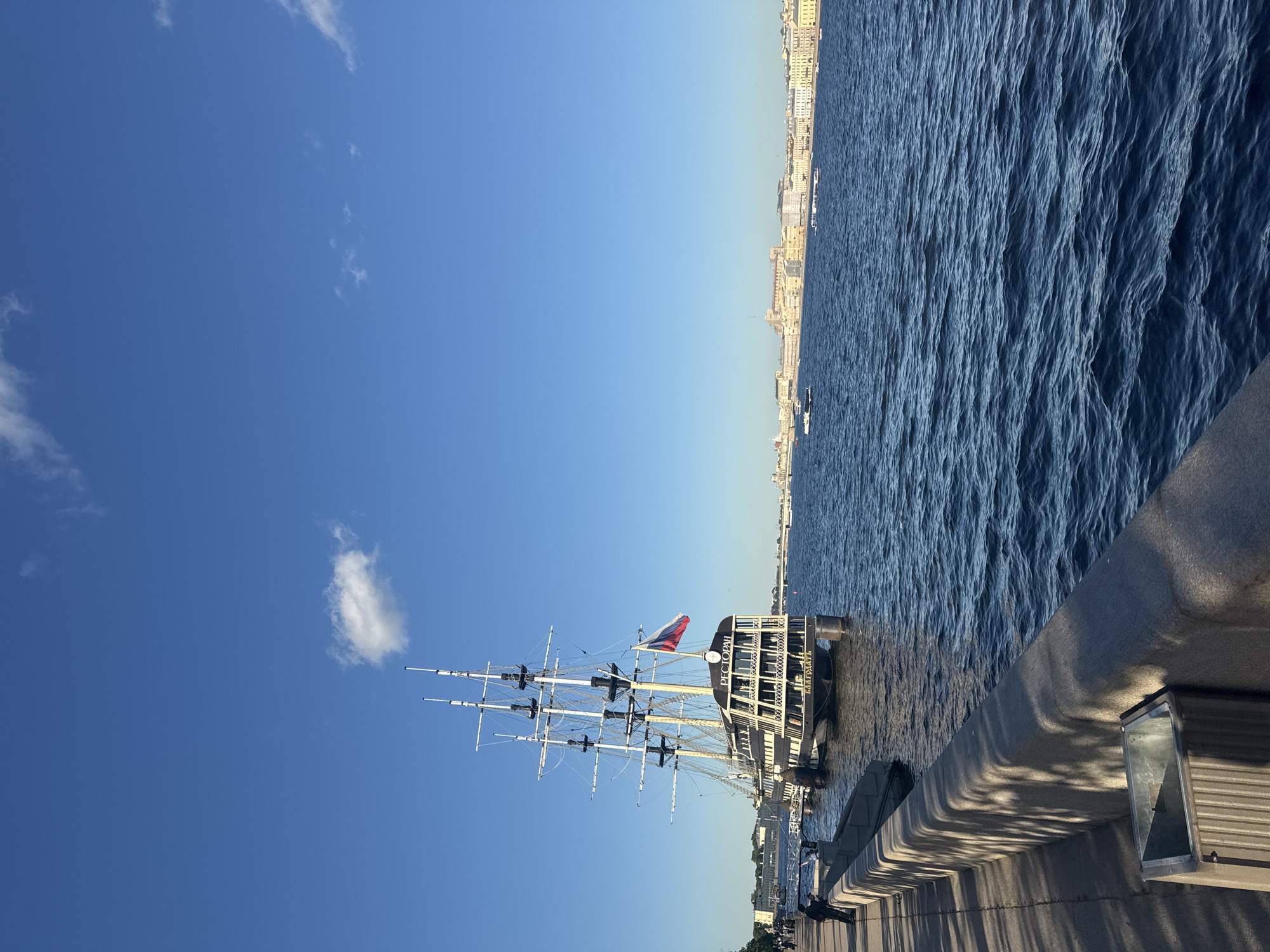 This is a photograph of a historic ship docked on the Neva River in Saint Petersburg, Russia. The ship, which appears to be a replica of a frigate, has three masts and is adorned with the Russian flag. The ship has the word "РЕСТОРАН" (Restaurant) visible on its side. In the background, the St. Petersburg skyline is visible with its characteristic classical architecture. The Neva River is calm with a few boats navigating the water. The foreground shows a stone embankment with a pedestrian walkway and a trash bin. A few people are walking along the embankment. The sky is a clear blue with a few scattered clouds, indicating a pleasant day. The image is captured in the afternoon, judging by the angle of the sun and the shadows cast on the embankment.