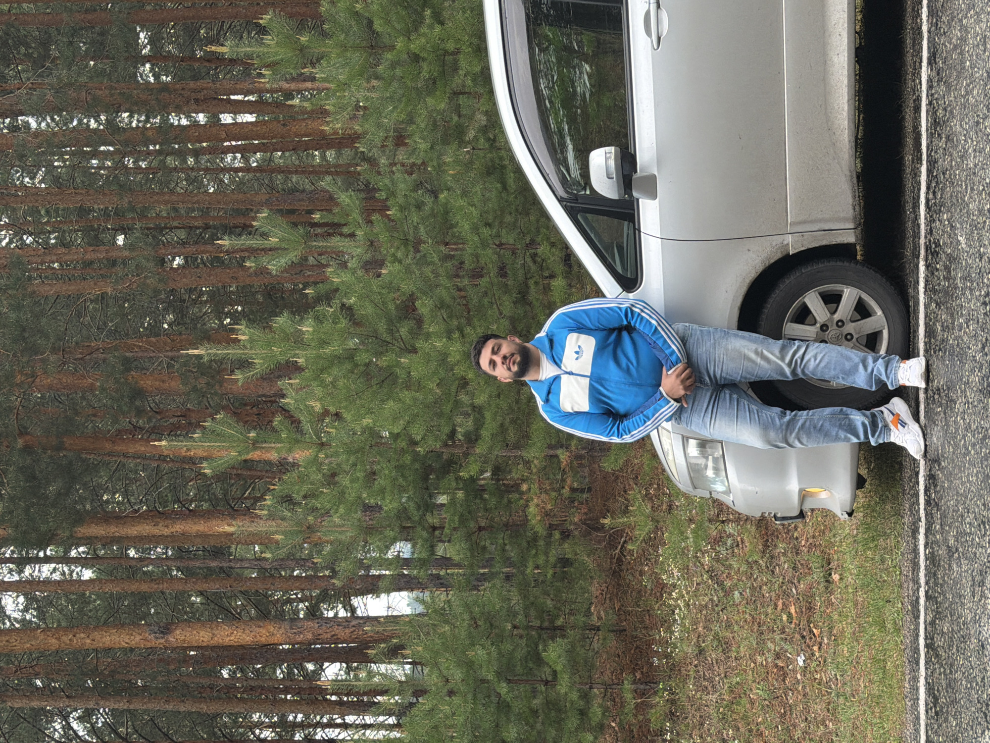 The image captures a man standing next to a silver van parked on the side of a road, with a dense pine forest visible in the background. The man is wearing a blue Adidas tracksuit and jeans. The forest is characterized by tall, straight pine trees. The location appears to be Сизая, Russia.