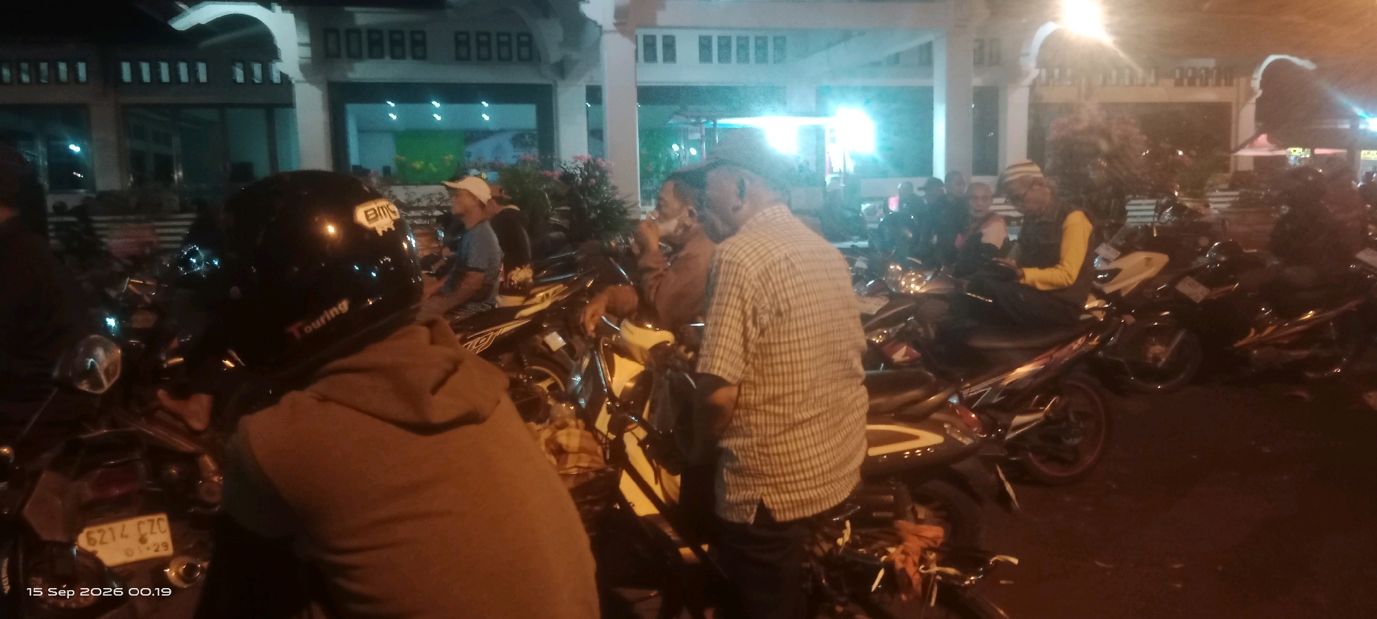 This is a nighttime photograph taken outdoors in the city of Klaten, Indonesia. The scene is crowded with motorcycles, suggesting a public gathering or event. In the foreground, a person wearing a black helmet with "BMC" and "Touring" visible is seated on a motorcycle. The license plate of this motorcycle reads "6214 CZC".
Several other people are visible, mostly sitting or standing near their motorcycles. They appear to be of various ages and are dressed in casual clothing, with some wearing head coverings. The background shows a building with illuminated windows and architectural details, likely a public facility or place of worship. The lighting is a mix of artificial sources, creating a somewhat dim and atmospheric environment. The ground appears to be paved, consistent with an outdoor public space in a city. The timestamp at the bottom of the image indicates it was taken on September 15, 2026, at 00:19.