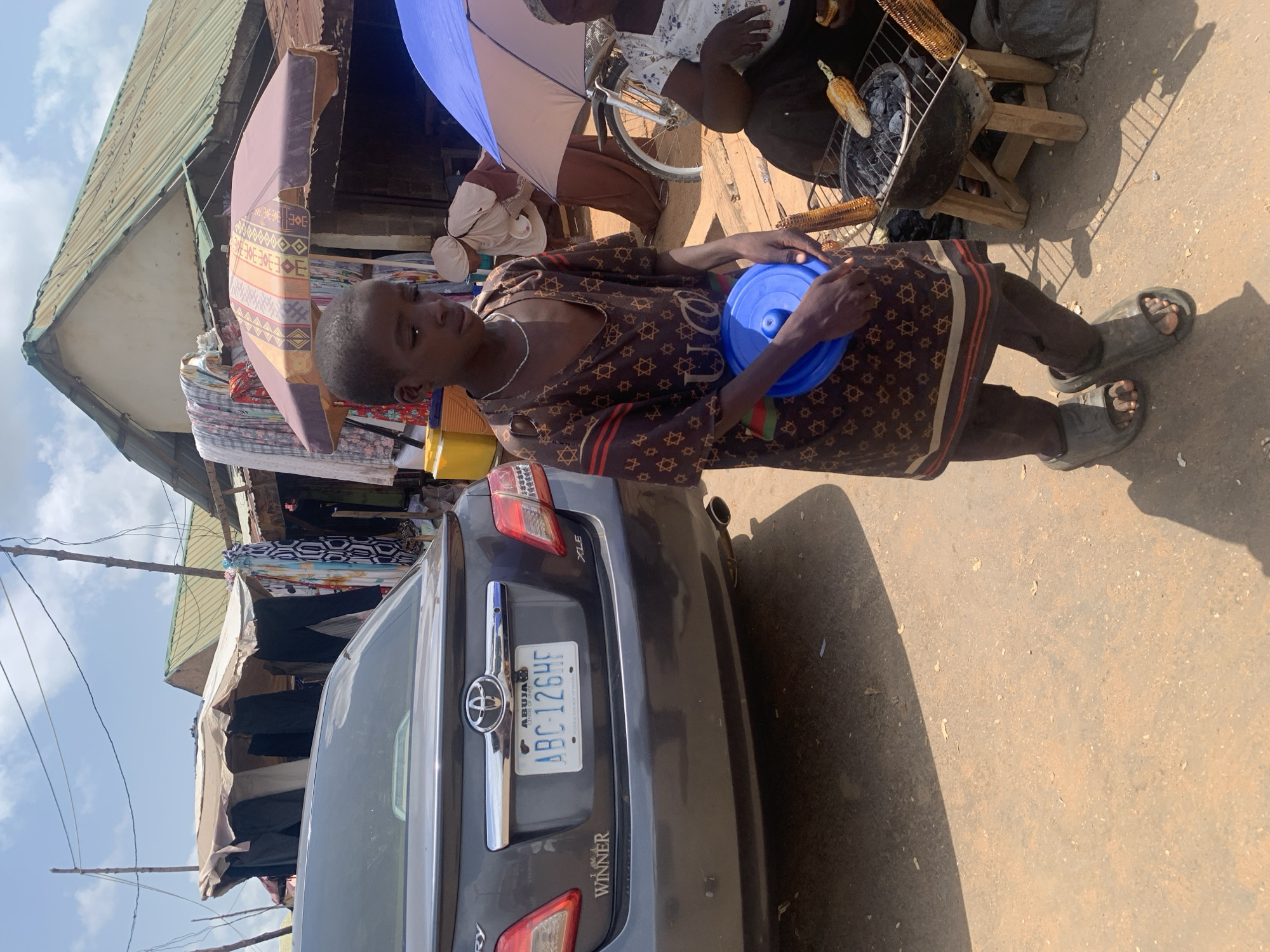 This is a street scene in Akwanga, Nigeria, on a sunny day. The foreground is dominated by a young boy, likely a vendor, wearing a brown patterned t-shirt and dark pants, holding a blue plastic lid. He is standing on a dirt road. To the right of the boy, there is a small charcoal grill with corn being roasted. Behind the boy, a woman is seated, partially obscured by an umbrella, attending to the grill. Further back, a dark grey Toyota Camry is parked, with its license plate visible: ABC-126HF. Adjacent to the car are market stalls with various textiles and goods hanging, covered by awnings. The overall environment suggests a busy open-air market setting.