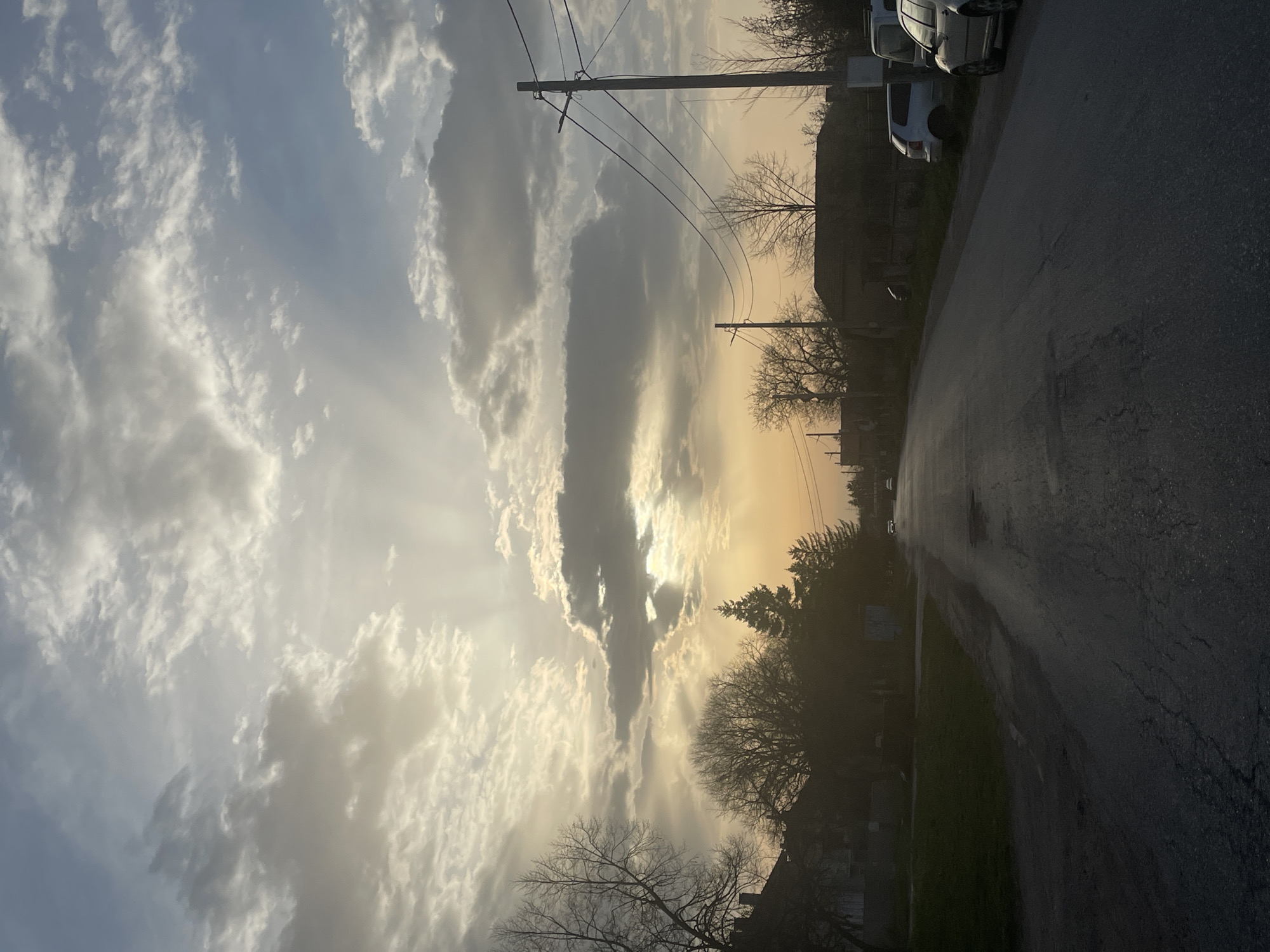 The image depicts a street scene, possibly in Aprilovo, Bulgaria, at sunset or sunrise. The sky is filled with clouds, with beams of sunlight breaking through. Power lines stretch across the sky, connecting poles on either side of the road. The road appears wet, reflecting light, and runs away into the distance. Buildings and trees line the road, silhouetted against the bright sky. Several cars are parked on the side of the road. There are no people visible in the scene.