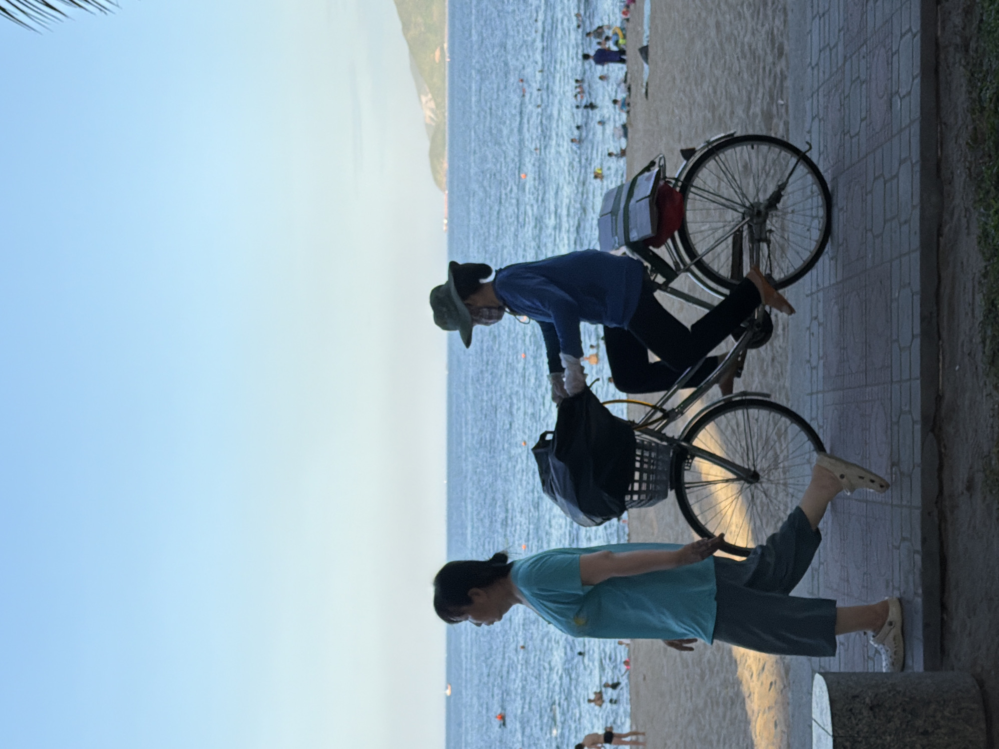 The image shows a beach scene in Nha Trang, Vietnam. In the foreground, a woman in a teal t-shirt and grey capri pants walks from left to right, facing away from the camera. To her right, a person is riding a bicycle, also moving from left to right. This person is wearing a blue long-sleeved shirt, dark pants, a wide-brimmed hat, and a face mask. The bicycle has a large black bag in the front basket and a red container on the back. The background features a sandy beach with several people swimming and relaxing in the ocean. The ocean is blue, and in the distance, hills are visible under a clear blue sky.