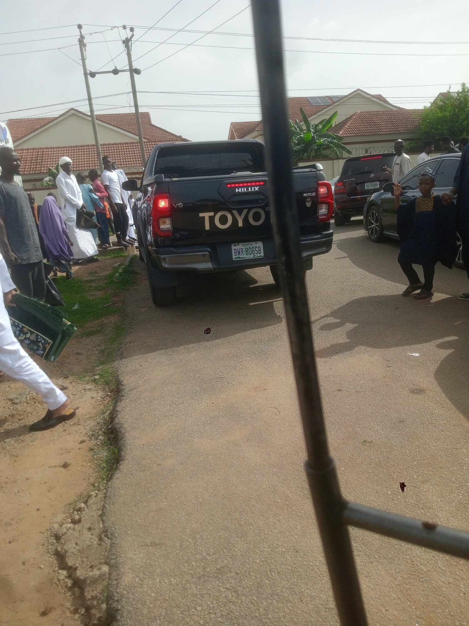 The image shows a street scene in Gwarinpa, Nigeria, with a black Toyota Hilux pickup truck parked in the middle of the road. The vehicle's license plate number is BWR-806SB. A group of people, some wearing traditional Muslim attire, are standing on the side of the road. Other vehicles, including a dark-colored Mercedes SUV and a dark-colored sedan, are also present. Houses with red-tiled roofs and solar panels are visible in the background. Utility poles with electrical wires are also seen. The foreground includes a portion of a metal structure obscuring part of the view. The road is paved and appears to be in a residential area.