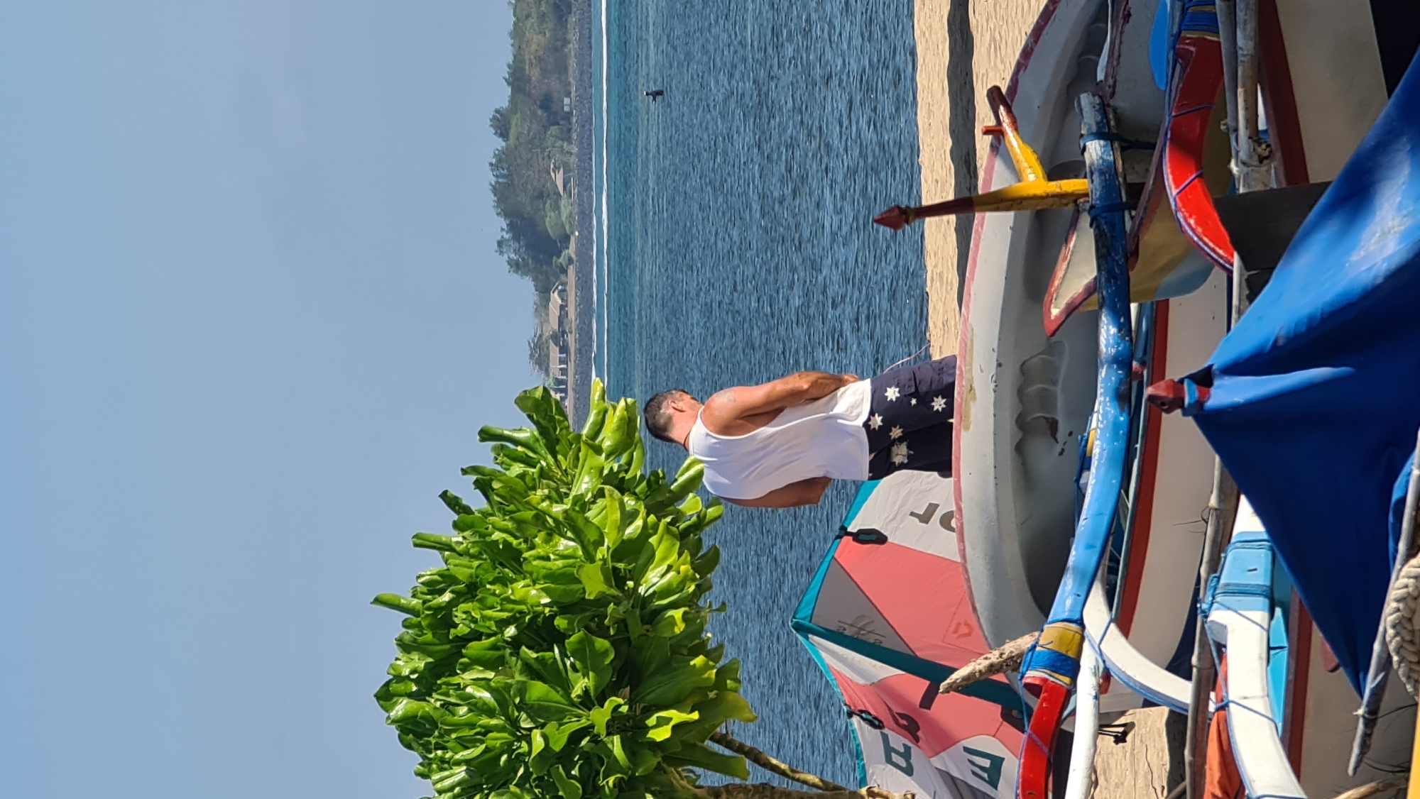 The image shows a man standing on a beach in Denpasar, Indonesia. He is wearing a white tank top and navy blue shorts with white stars. He is urinating. In the background, there is the ocean with a few boats on the shore. There is also a tree with green leaves and some buildings in the distance. The sky is clear and blue.