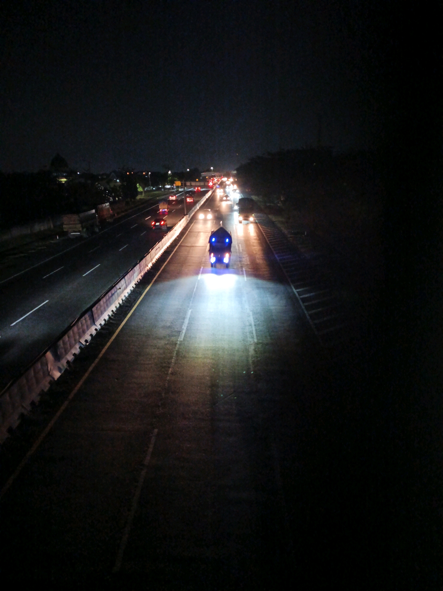 An elevated, wide-angle shot captures a highway at night in Kludan, Indonesia, with traffic flowing in both directions. The foreground is dominated by the dark asphalt of the highway, illuminated by the bright headlights of oncoming vehicles and the taillights of those receding. A lone motorcycle with bright blue and white lights is in the center of the frame, its lights casting a strong glow on the road ahead. The motorcycle appears to be moving away from the viewer. On the left side of the highway, there are several lanes with vehicles visible, their headlights and taillights creating streaks of light. A concrete barrier with a yellow stripe separates the lanes. On the right side of the highway, the scene is darker, with trees and foliage lining the road. The sky is black, indicating it is nighttime.