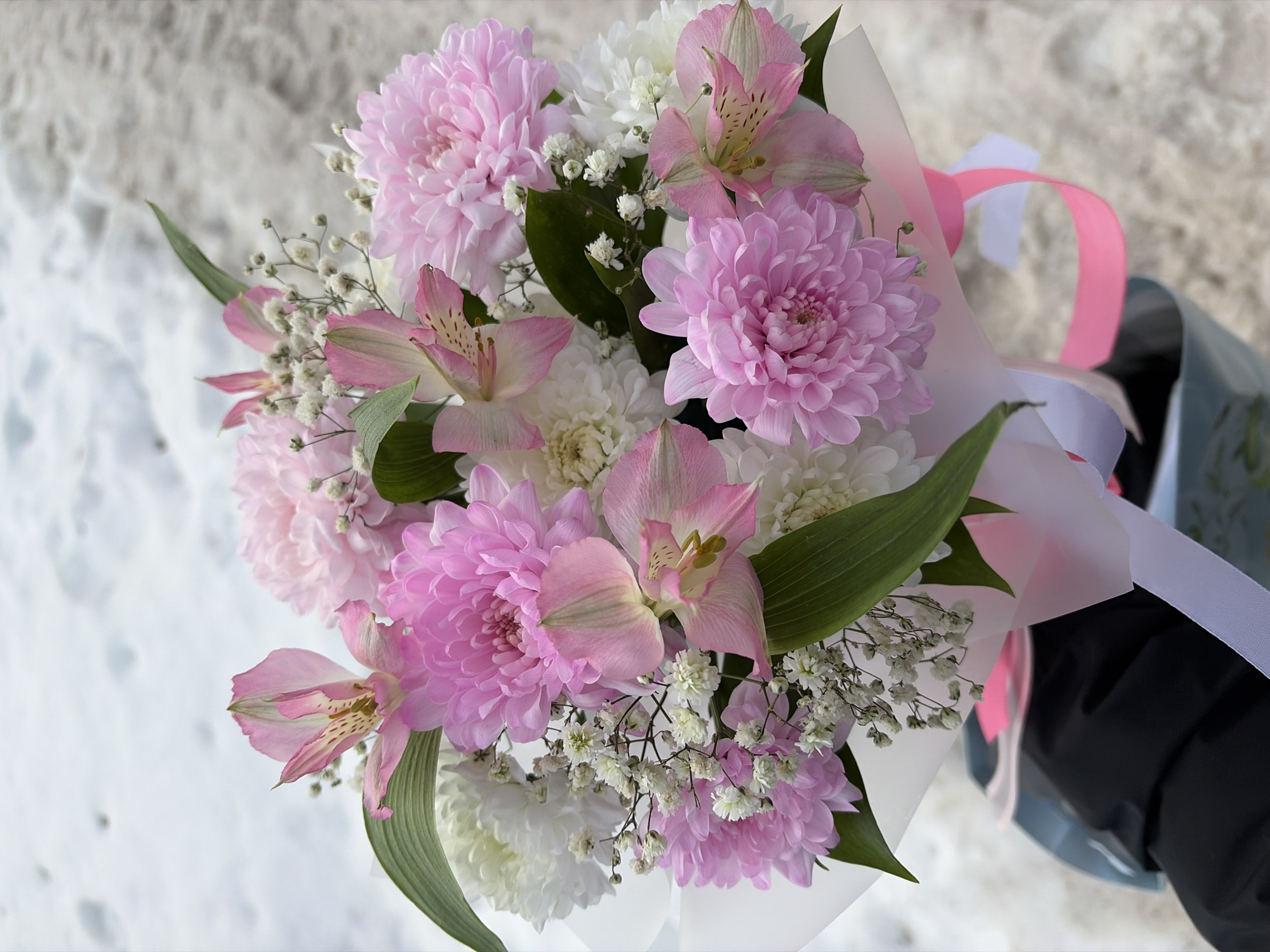 This image displays a floral bouquet held by an individual outdoors, set against a snow-covered ground in Chaikovsky, Russia. The bouquet consists of several densely petaled pink chrysanthemums, white chrysanthemums, and pink alstroemeria flowers, which feature characteristic dark speckling on their inner petals. Small clusters of white baby's breath (Gypsophila) are interspersed throughout, along with various dark green foliage leaves. The bouquet is wrapped in white, translucent material and secured at the base with interwoven pink and white ribbons. A partial view of a dark-colored sleeve, consistent with a winter jacket or coat, is visible on the person holding the bouquet. Below the bouquet, a segment of a light blue-green container or bag, showing subtle patterned graphics, is visible. The background consists of textured, compacted snow, suggesting an outdoor, possibly urban or semi-urban, environment. No specific landmarks are discernible to precisely identify the location within Chaikovsky.