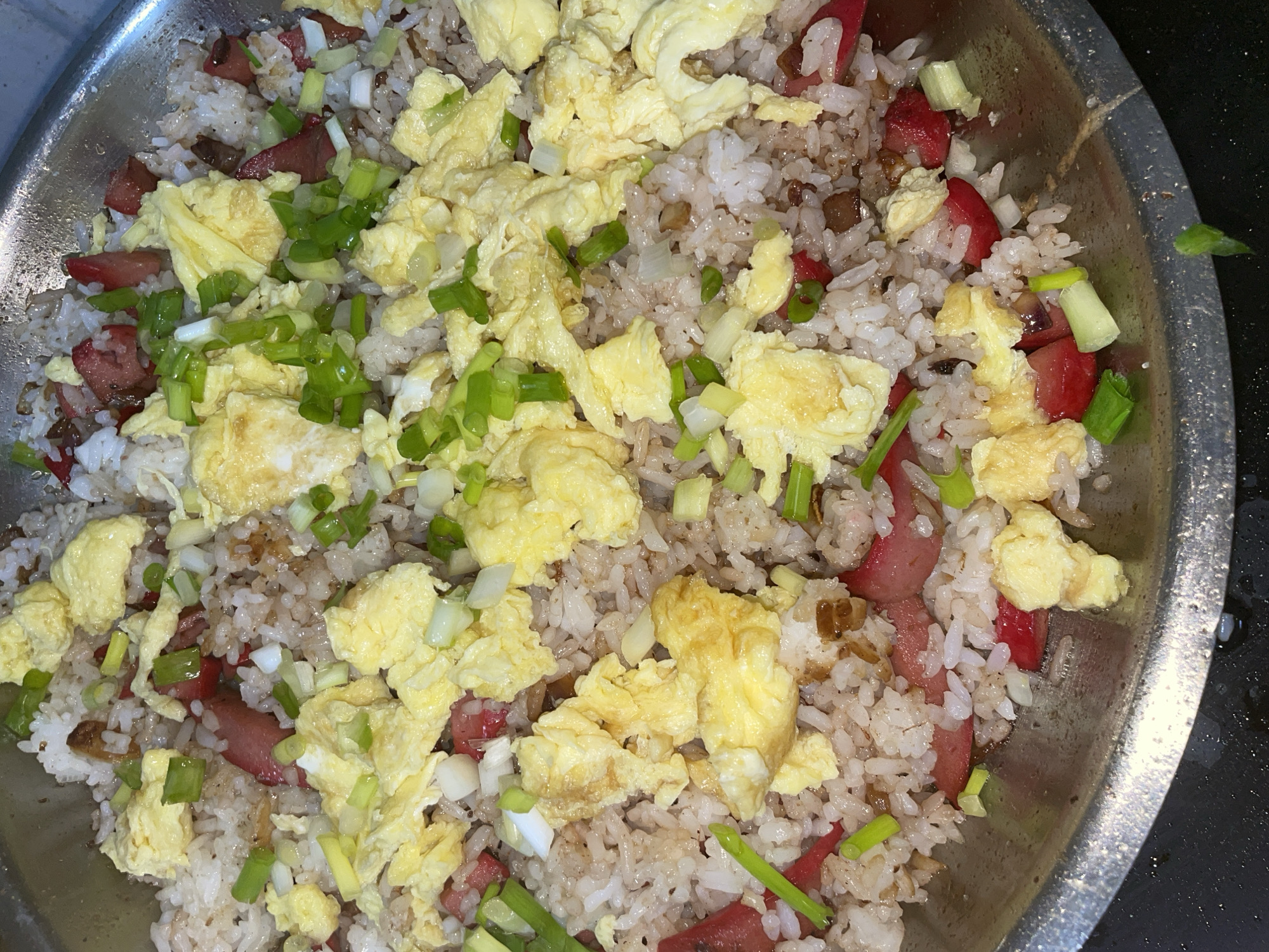 This is a close-up, overhead shot of a large bowl of fried rice. The bowl appears to be made of metal and is filled to the brim with the dish. The fried rice contains white rice, scrambled eggs, sliced red sausage, and chopped green onions. Some darker, possibly savory bits are also interspersed throughout the rice. The lighting suggests it is indoors, possibly in a home kitchen, given the slightly messy appearance of the bowl's rim and the dark, textured surface it rests on, which could be a countertop. There is no visible text in the image. The meal appears to be freshly prepared and ready to be served, with the ingredients mixed together in a hearty portion. The overall impression is of a comforting and familiar homemade dish. The location context of Davao City, Philippines, suggests this might be a local variation of fried rice, perhaps incorporating common Filipino ingredients or cooking styles.