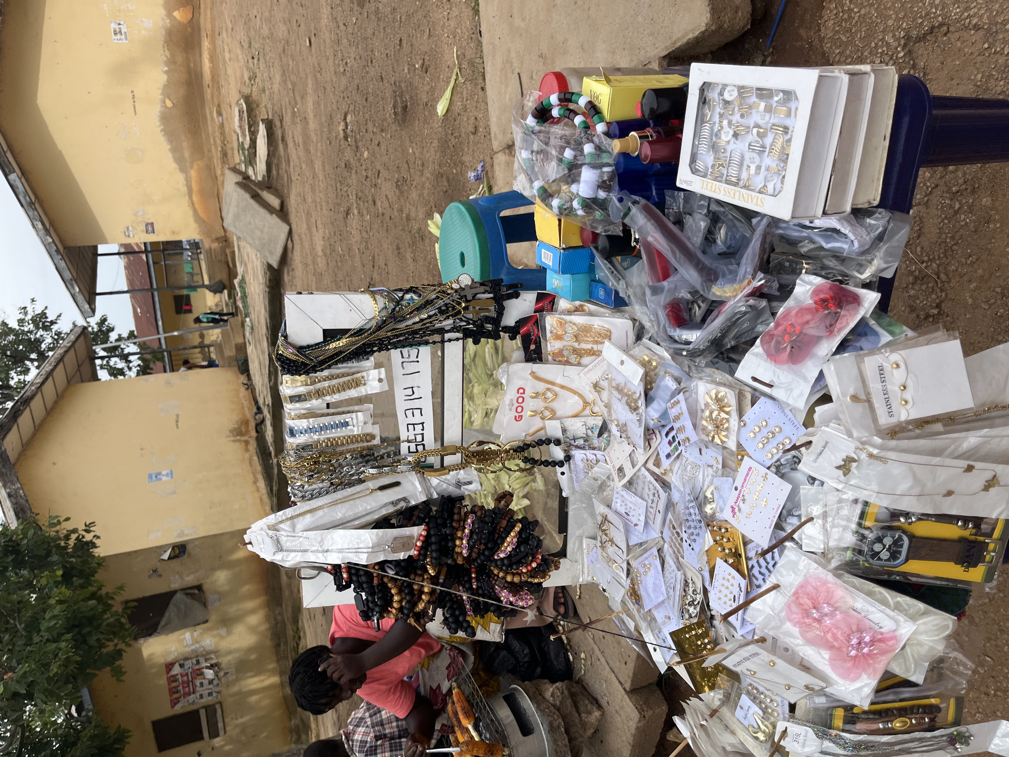 An outdoor market scene in Jos, Nigeria features a stall displaying various accessories. A person wearing a pink t-shirt is seen to the left, with a small grill in front of them cooking corn. The stall is laden with an assortment of items including necklaces, bracelets, earrings, watches, and rings, all packaged and displayed for sale. A blue stool is placed behind the stall. The background shows two yellow buildings with a somewhat weathered appearance and a dusty, unpaved ground.