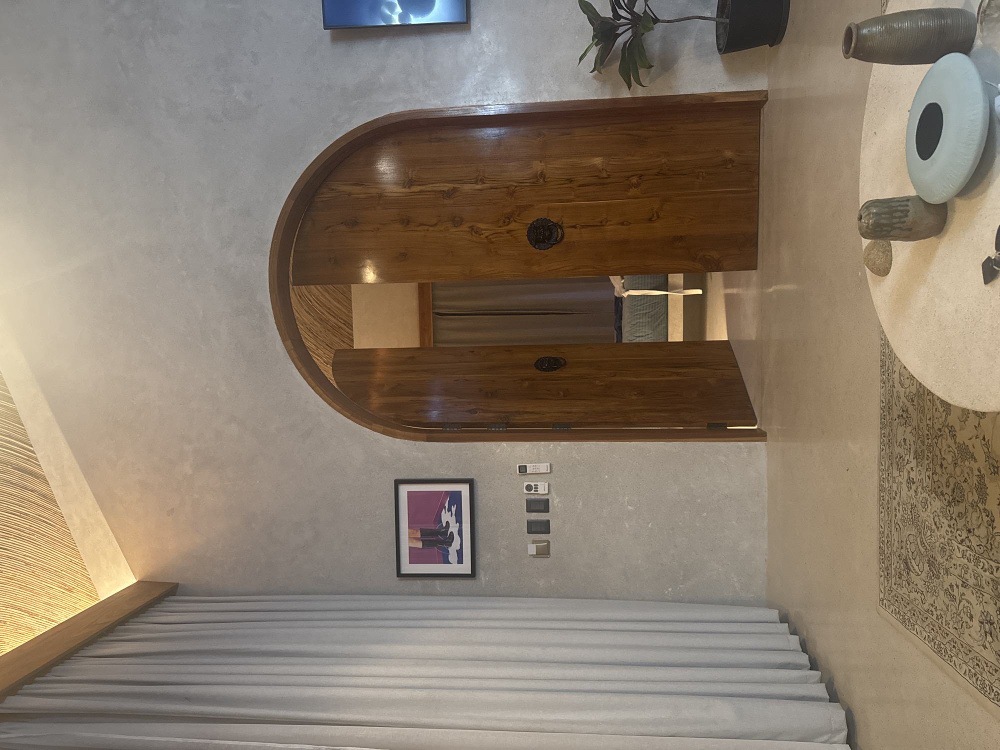An interior view depicts a modern room, identified as being in El Nido, Philippines. The space features light grey textured walls and a vaulted ceiling, partially covered with a light brown, woven natural material. A recessed light strip runs along a wooden beam near the ceiling. On the left, floor-to-ceiling light-colored curtains obscure a window. A framed artwork, depicting legs in black boots standing in a white puddle against a purple background, hangs on the left wall. Below it, a series of wall-mounted controls includes three light switches and two remote controls.
Centrally positioned is an arched, double wooden door with a natural wood finish and dark lion-head door knockers. The right door is slightly ajar, revealing a glimpse of an adjoining room with a bed featuring a light blue comforter and a similar woven ceiling. To the right of the door, a dark-screen television displaying a light-colored, cloud-like image is mounted on the wall. Below the television, a potted plant with dark green leaves rests in a black pot on the floor.
The floor is a light-colored, smooth surface, possibly polished concrete. A patterned beige and brown rug with an intricate design is partially visible in the foreground. A round, light-colored coffee table occupies the lower right foreground, displaying various decorative items: a light brown cylindrical vase, a small irregular stone, a flat light blue oval object with a dark center, a taller dark brown ribbed vase, and a set of keys. No individuals are present in the scene.