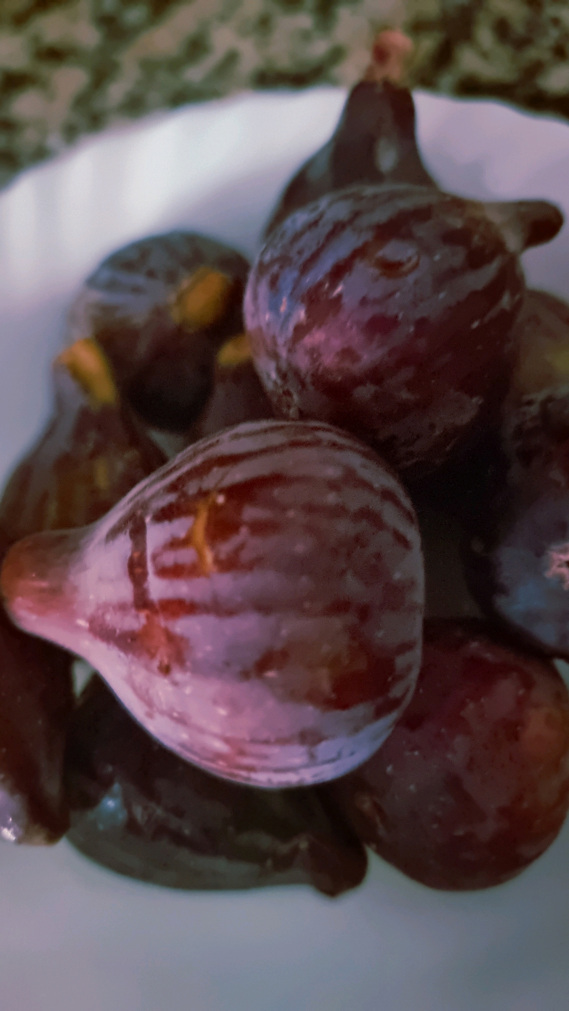 A close-up, slightly out-of-focus shot shows a pile of ripe figs in a white bowl. The figs are dark purple, with visible stripes and a shiny skin. One fig in the foreground is particularly prominent, showing its textured surface and a small stem. The background is blurred, with a mottled green and brown pattern, likely a tablecloth or countertop. The lighting is dim, casting subtle reflections on the figs. The setting is domestic, with no obvious indicators of the specific location within Sant Joan Les Fonts, Spain.