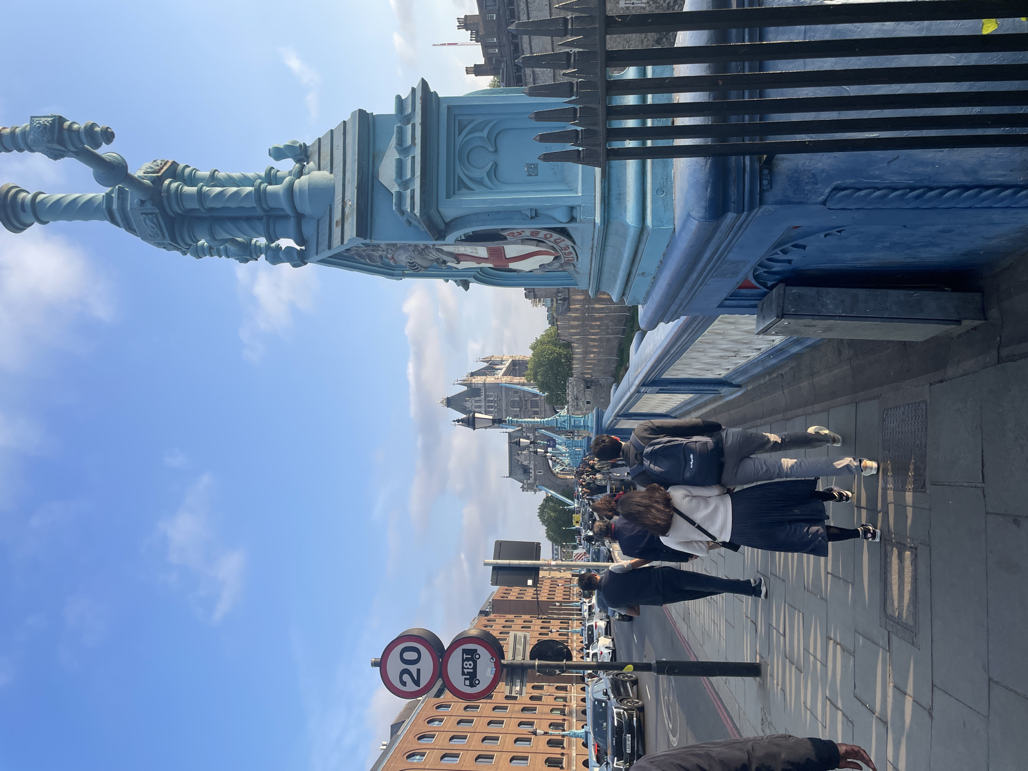 The image depicts a view from the Tower Bridge in London, United Kingdom. Pedestrians, including a group walking close together, are present on the bridge's walkway. The bridge's distinctive blue painted ironwork is prominent in the foreground. A 20 mph speed limit sign and an 18T weight restriction sign are visible beside a brick building. In the background, the Tower of London can be seen. The sky is blue with scattered clouds.
