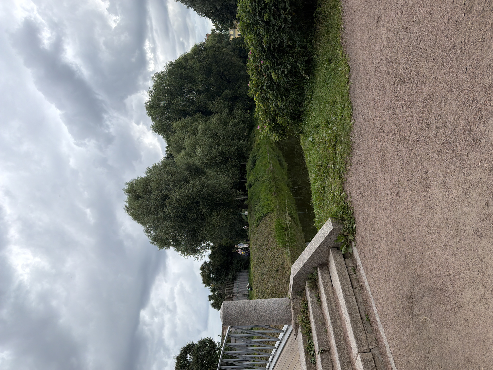 The image depicts a park scene with a canal and lush green trees under a cloudy sky. In the foreground, a sandy path leads to a stone staircase and a granite pillar. A metal railing of a bridge is visible on the left. Across the canal, people are seen walking along the grassy bank. The environment suggests a tranquil, outdoor setting within a city, possibly a public garden or park. The architecture and general ambiance are consistent with locations in Saint Petersburg, Russia.