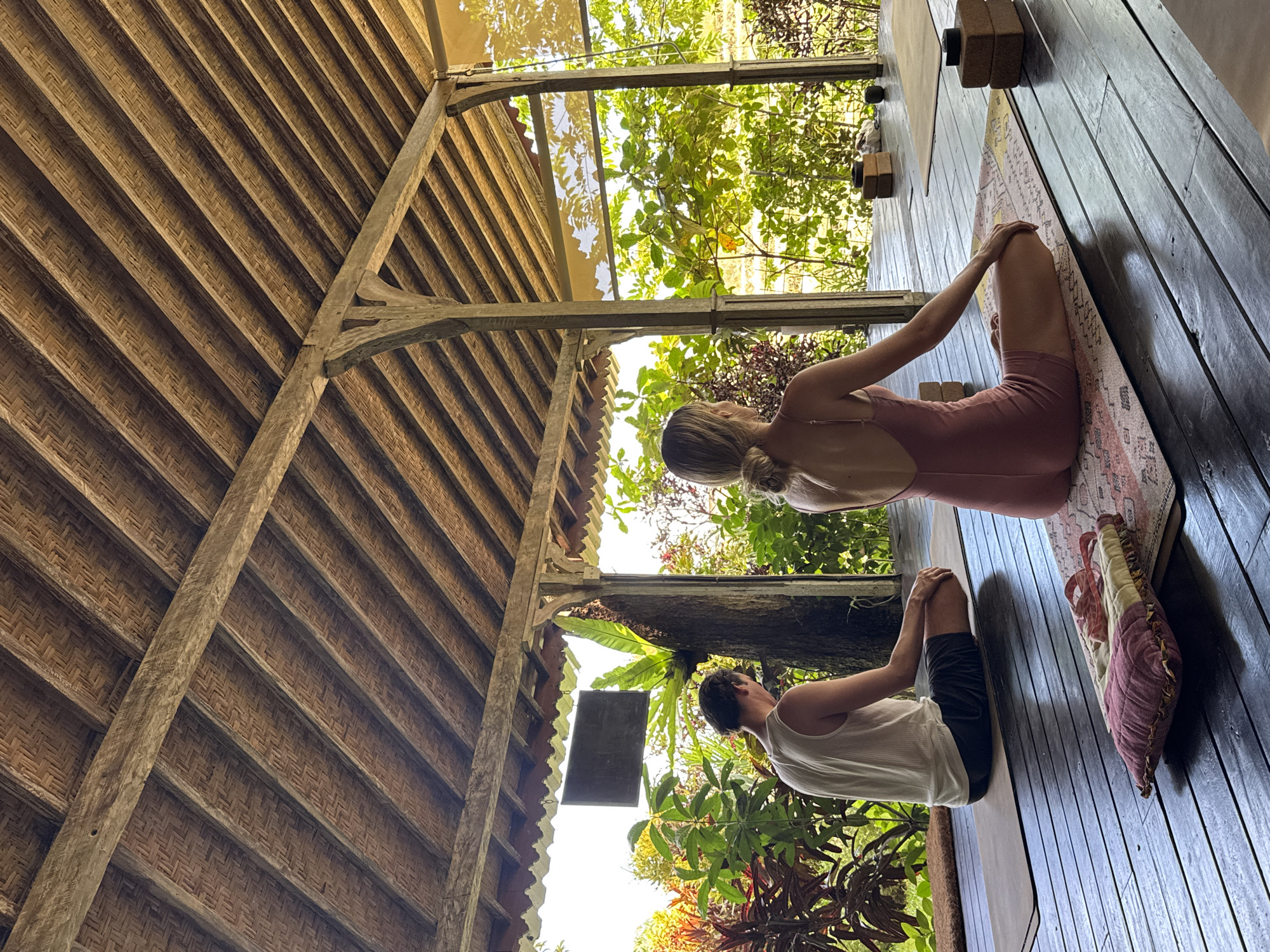 A medium shot shows two people on a yoga mat in a rustic, open-air structure, likely in Mengwi, Indonesia. The woman in the foreground is wearing a pink bodysuit and sitting in a butterfly pose. The man behind her is wearing a white tank top and black shorts, also in a butterfly pose. The structure has a thatched roof and wooden beams. Lush greenery surrounds the structure. A yoga mat is on a wooden floor. Yoga blocks are visible near the woman. A pillow is visible on the floor next to the yoga mat.