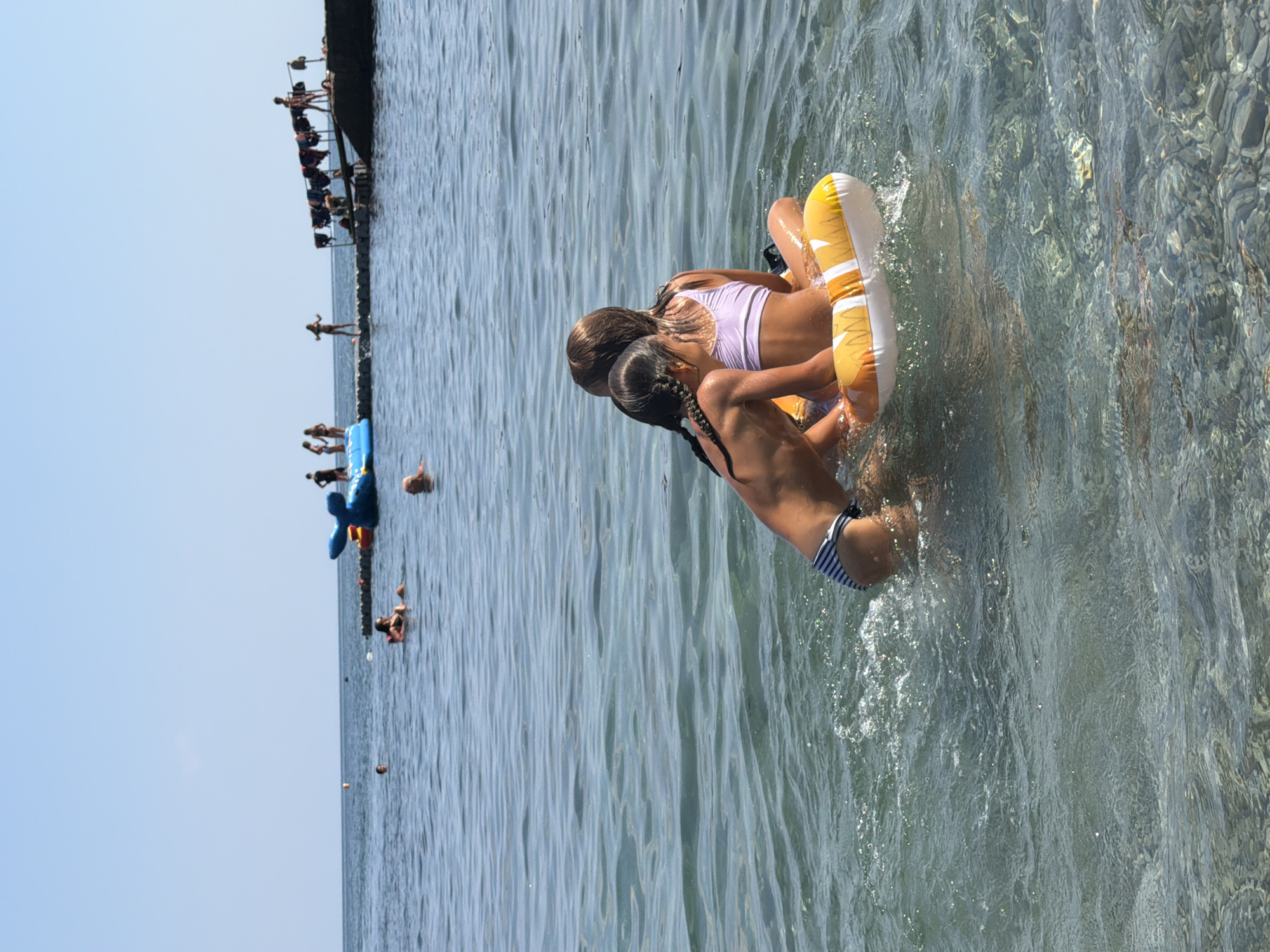 In this image, two girls are playing in the sea on an inflatable ring. The girl in front is wearing a white top and has her hair in a braid. The girl behind her is wearing a purple swimsuit. They are both in the water, with the inflatable ring between them. The water is clear and blue, and there are ripples on the surface. In the background, there is a pier with several people on it, and some inflatable toys in the water. The sky is clear and blue, suggesting it is a sunny day. The overall scene depicts a leisurely day at the beach.