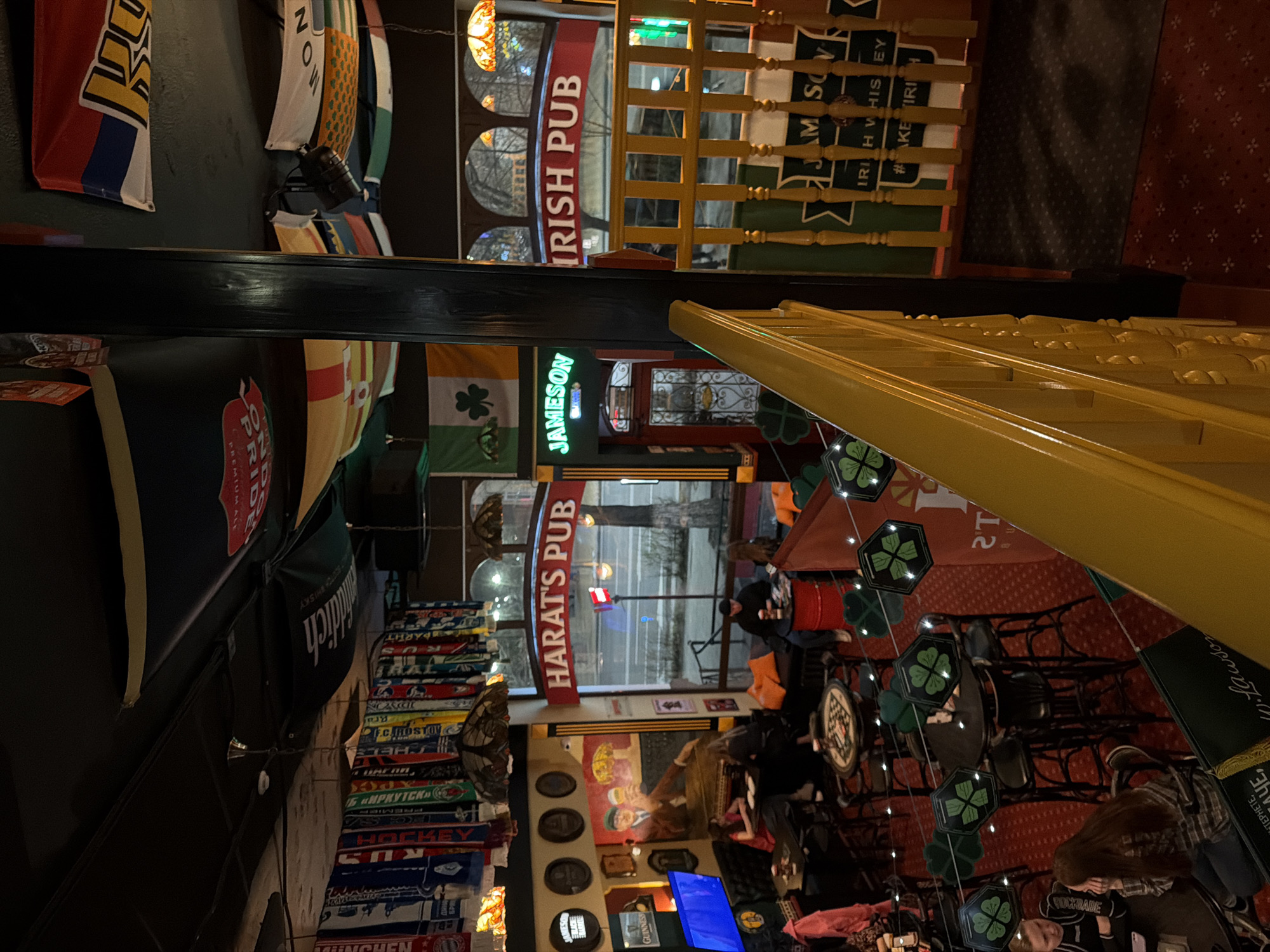 Here is a detailed description of the image:
The image depicts the interior of an establishment identified as an Irish pub, named "Harat's Pub", and "Irish Pub" in Rostov-on-Don, Russia. The orientation of the image is tilted. Several patrons are seated at tables and chairs within the pub. Various decorations are visible, including shamrock themed items. Banners and flags with possibly hockey related logos line one wall. Signs advertising "Jameson" and "Guinness" are also noticeable. There are wooden stairs.