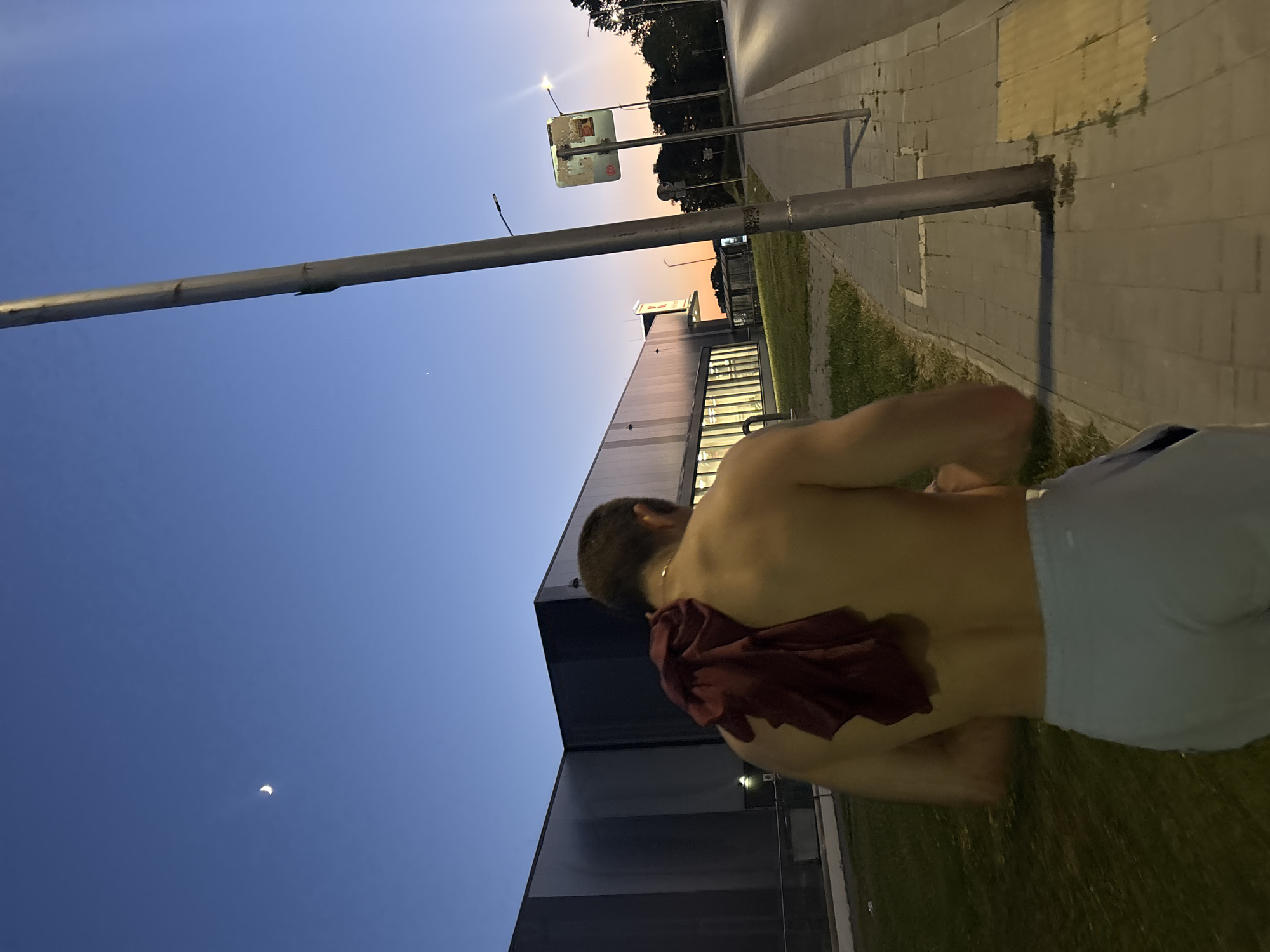 A shirtless person with a maroon towel draped over their shoulders walks away from the camera on a sidewalk. The person is wearing light blue athletic shorts. They are walking down a sidewalk next to a road and a building that appears to be a large commercial establishment, possibly a supermarket or a warehouse. The sky is a deep blue, with a crescent moon visible on the left side of the frame. The setting appears to be dusk, with streetlights beginning to illuminate the scene. The location is the area in front of Kaufland Varna, which is located on Vladislav Varnenchik Blvd. in Varna, Bulgaria.