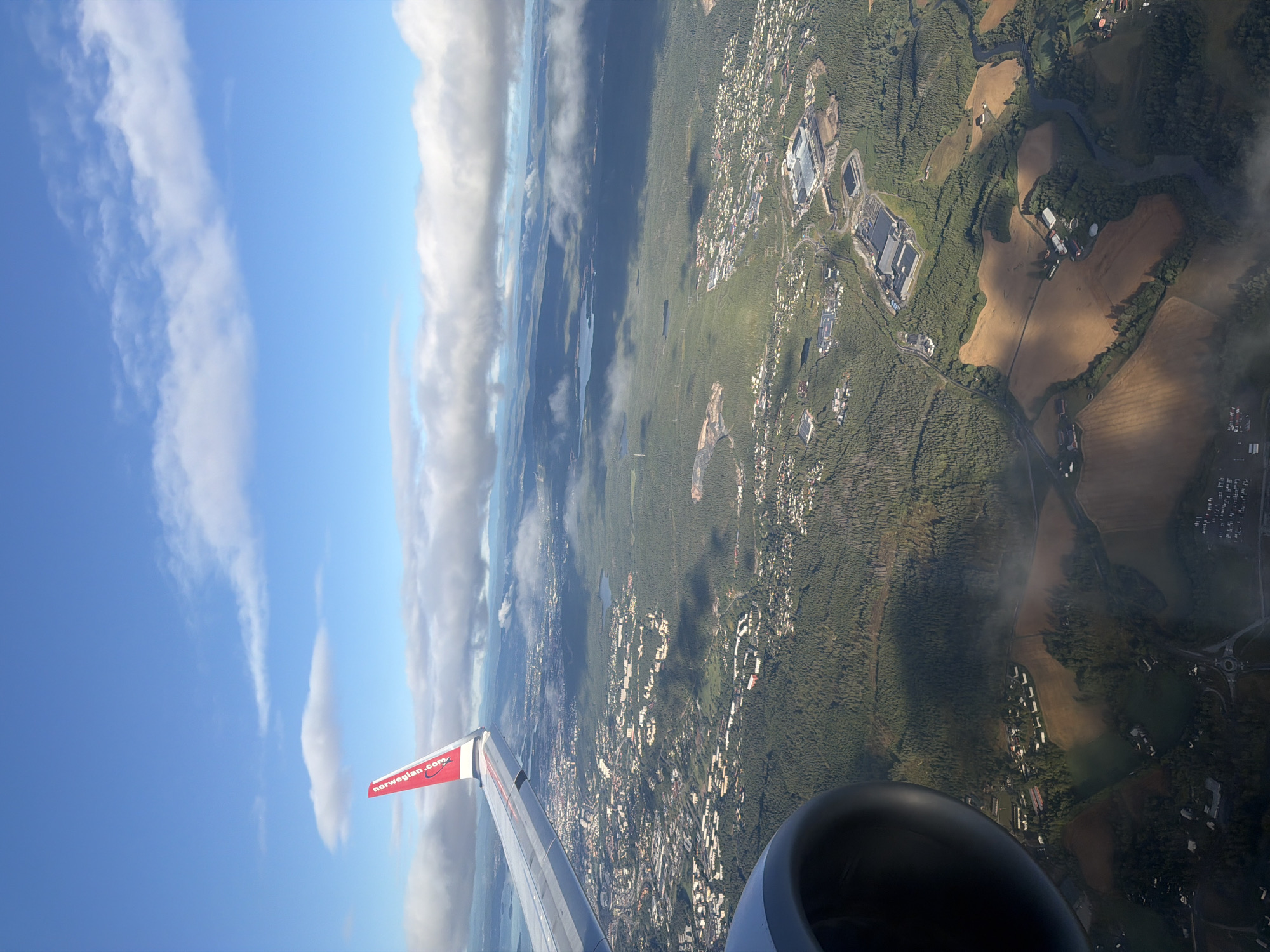 An aerial view captured from an airplane window shows a sprawling landscape under a partly cloudy blue sky. The foreground features a portion of an airplane engine and wing, with the distinctive red logo and lettering "norwegian.com" clearly visible on the wing.
The midground and background reveal a densely wooded area interspersed with patches of cultivated fields and a scattering of buildings, indicating a suburban or semi-rural environment. The terrain appears somewhat hilly, with bodies of water visible in the distance. Shadows cast by clouds create dramatic contrasts across the landscape.
Specific landmarks in Lillestrøm, Norway, are identifiable:
* **Øyerfjellet Nature Reserve:** The extensive forested areas visible in the image likely correspond to the natural reserve.
* **Skedsmohallen:** A large, modern building complex with a light-colored roof, situated within the forested area and near a road, is likely Skedsmohallen, a sports and events venue.
* **Sofiemyr:** A residential area characterized by clusters of houses, some with white facades, is visible within the wooded terrain.
* **Flax:** A large industrial building with a grey roof, located near the center of the frame, is identifiable as Flax, a distribution center.
* **Nordre Fjellmyr:** A residential area with numerous houses is visible to the left of the image.
* **The Nitelva River:** A winding body of water, likely the Nitelva River, meanders through the landscape.
* **Rotnes:** A circular intersection on a road is visible in the lower part of the image, indicating a junction in the urbanized area.
