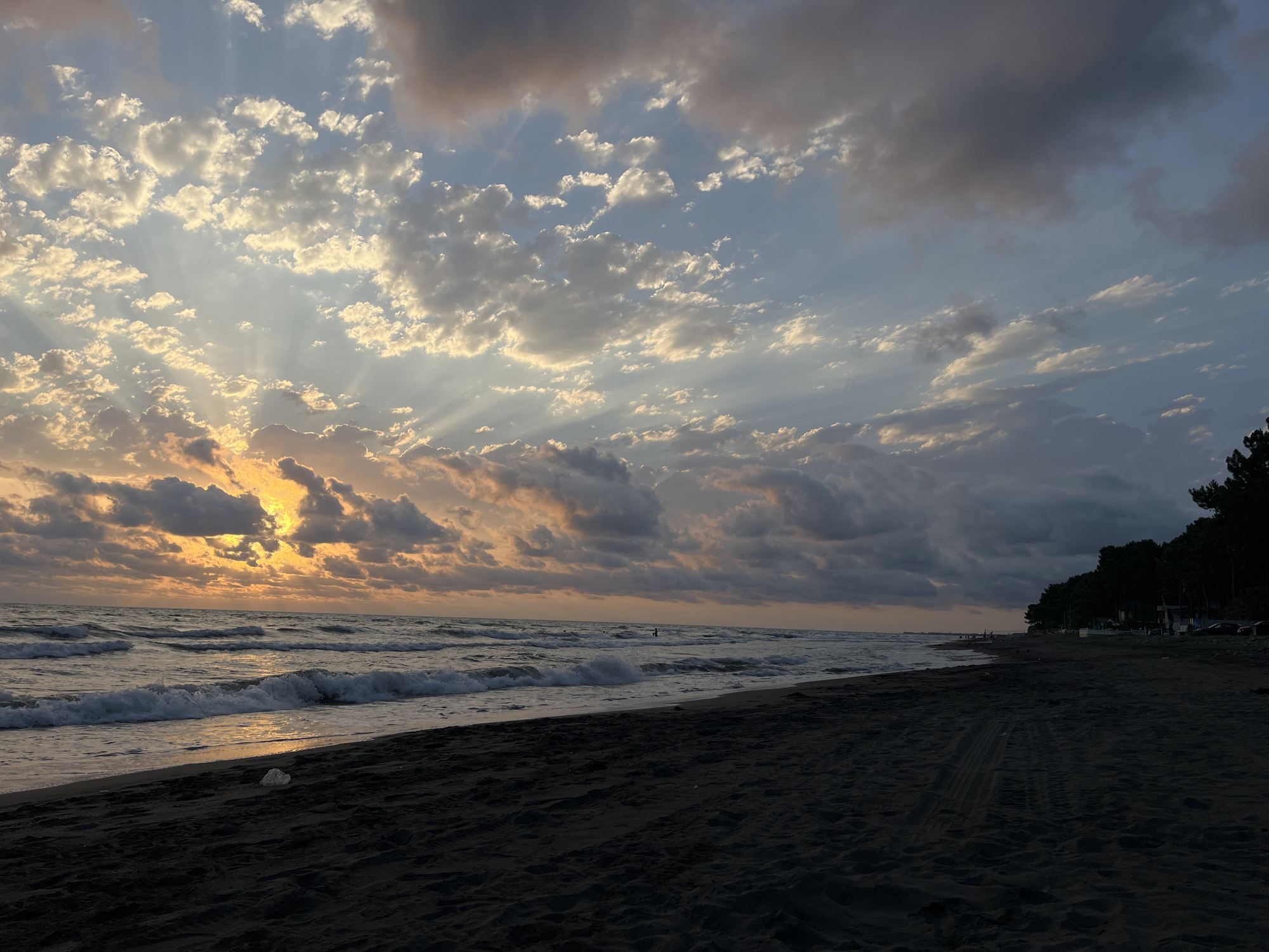 This is a photograph of a beach at sunset. The sky is filled with dramatic clouds, with rays of sunlight breaking through. The sun itself is partially obscured by clouds, casting a warm orange glow on the scene. The ocean is a deep blue, with waves gently rolling onto the dark sand beach. In the distance, there is a line of trees and some buildings, suggesting a coastal town or resort. There are tire tracks on the sand, indicating vehicle access. A few people are visible on the beach and in the water, but they are small in the frame and their actions are not discernible. The overall mood is peaceful and serene, with the natural beauty of the sunset taking center stage.