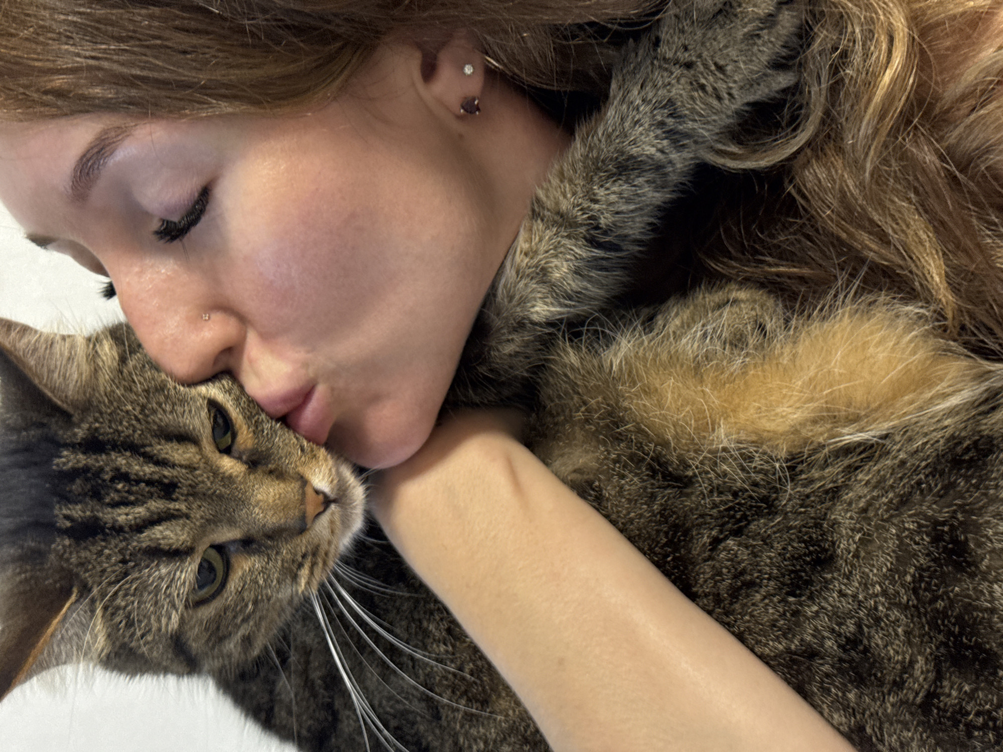 The image shows a close-up of a person kissing a tabby cat. The person's eyes are closed, and they are leaning in to kiss the cat on its cheek. The cat appears to be looking at the camera with a slightly annoyed expression. The person has long, auburn hair and is wearing a furry coat. The background is a plain white wall. There is no indication of the city of Chaikovsky, Russia in the image.