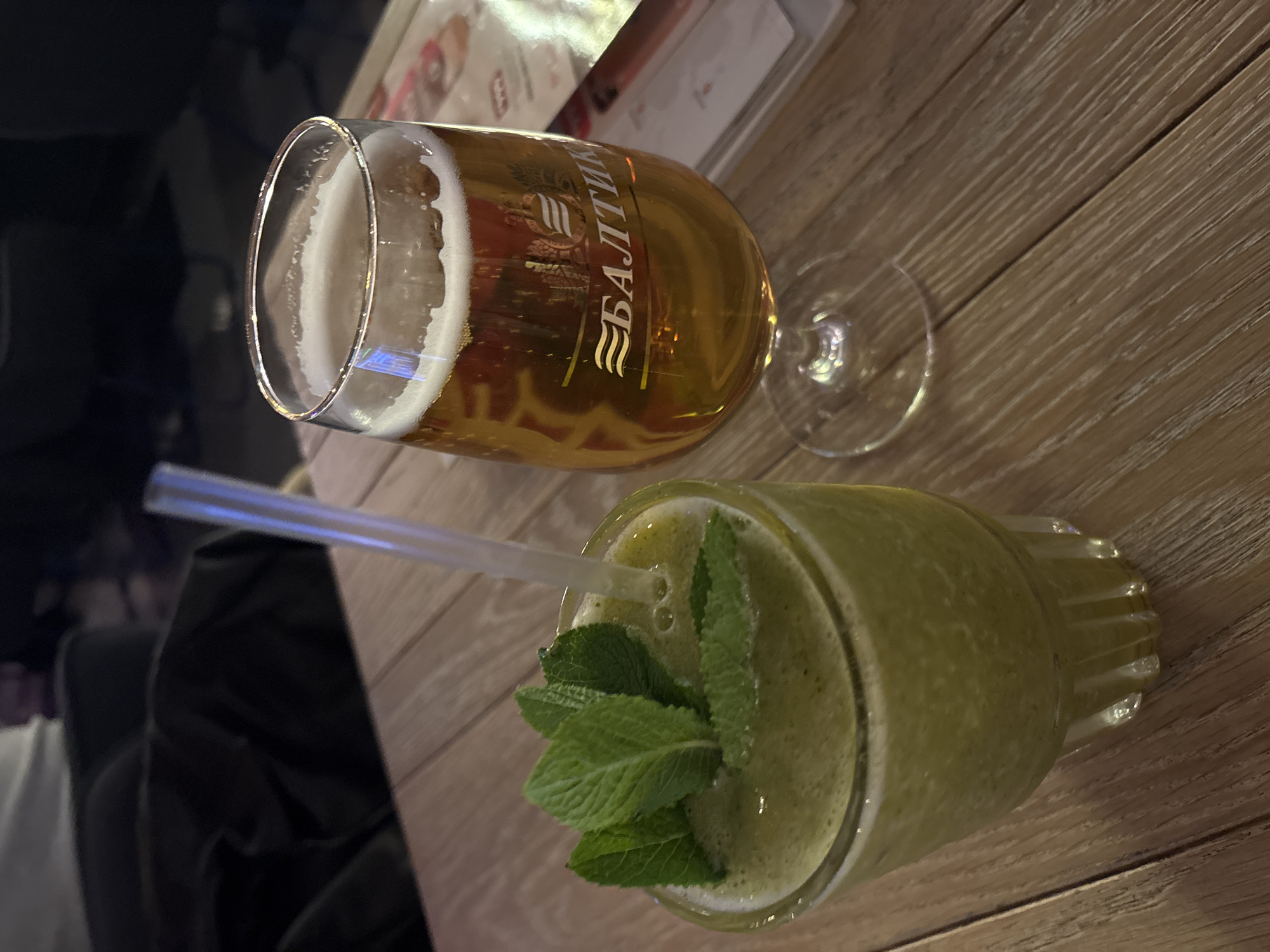 The image, taken in Vsevolozhsk, Russia, captures two beverages resting on a light brown wooden table, viewed from a slightly elevated perspective. The lighting is dim, suggesting an indoor setting like a cafe, restaurant, or bar, likely in the evening.
On the left, a tall, stemmed glass holds a golden-hued beer with a substantial head of white foam. The side of the beer glass clearly displays the brand name "БАЛТИКА" (BALTIKA) in white Cyrillic letters, accompanied by a logo featuring three wavy lines and an emblem.
To the right, a wider glass contains a vibrant green smoothie or juice, garnished with several fresh mint leaves floating on its surface. A transparent straw is positioned in the center of the green drink, which appears thick and slightly frothy.
The table features a noticeable light wood grain pattern. In the soft-focus background, indistinct shapes and reflections are visible, including what appears to be a menu or piece of paper and dark, blurred elements that could be furniture or other patrons.