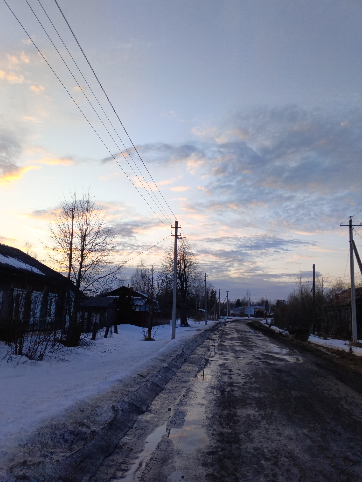 An outdoor scene in Красный Маяк, Russia, depicts a rural or semi-rural street during dusk or dawn. The unpaved road, appearing wet and muddy with numerous puddles, extends into the distance, showing signs of melting snow. Dirty snowbanks line both sides of the road. On the left, a dark, aged wooden house with a sloped roof is partially visible, flanked by bare deciduous trees and bushes. Utility poles with multiple overhead wires are present on both sides of the road, extending towards the horizon. The sky, occupying the upper half of the frame, displays a gradient from soft orange and yellow hues on the left horizon to light blue and grey overhead, with scattered stratocumulus clouds. In the background, more indistinct houses and bare trees line the curving road. No individuals are visible, and no actions or interactions are depicted.