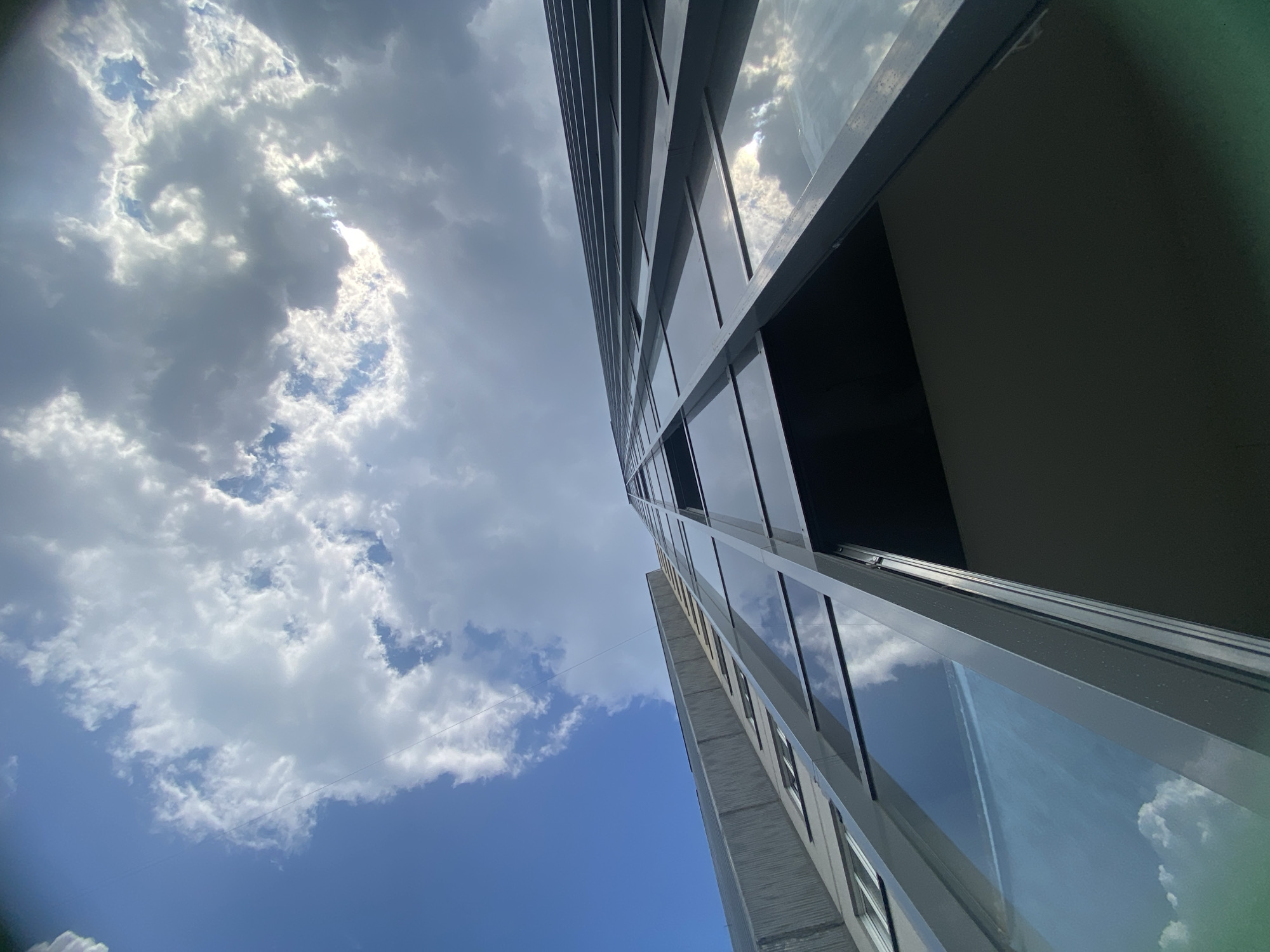 This is a low-angle, upward-facing shot of a modern building with a glass facade, set against a sky filled with dramatic clouds. The building's exterior is characterized by dark, reflective panels and a series of horizontal frames. The perspective emphasizes the height and scale of the structure. The sky is a dynamic mix of bright blue patches and large, billowing grey and white clouds, with sunlight filtering through in places. A thin, dark line, possibly a wire, is visible stretching across the sky. The building's reflective windows mirror the clouds and sky, adding to the visual complexity of the scene. The image appears to be taken from ground level, looking up at the building's edge and the sky above. The overall impression is one of urban architecture interacting with a dramatic natural sky. There are no people or animals visible in the image. This image is taken in the city of Novosibirsk, Russia.